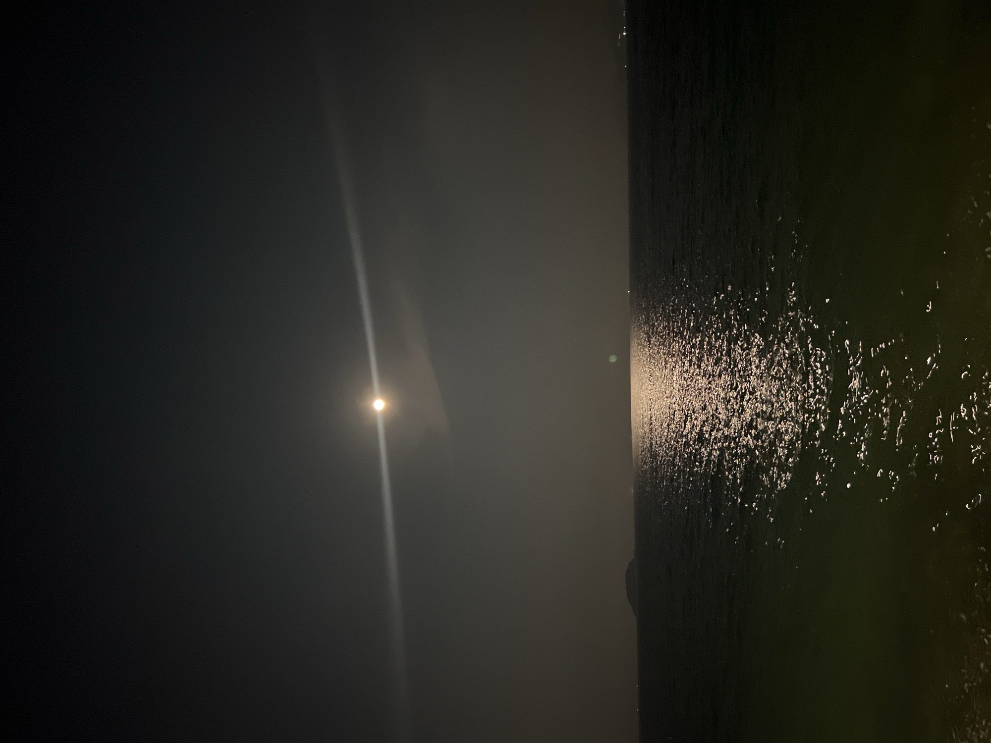 The image captures a night scene in Bắc Nha Trang, Vietnam. The moon is visible in the upper left, casting a bright beam downwards. To the right, dark water reflects the moonlight. The environment is dark and potentially foggy or hazy.