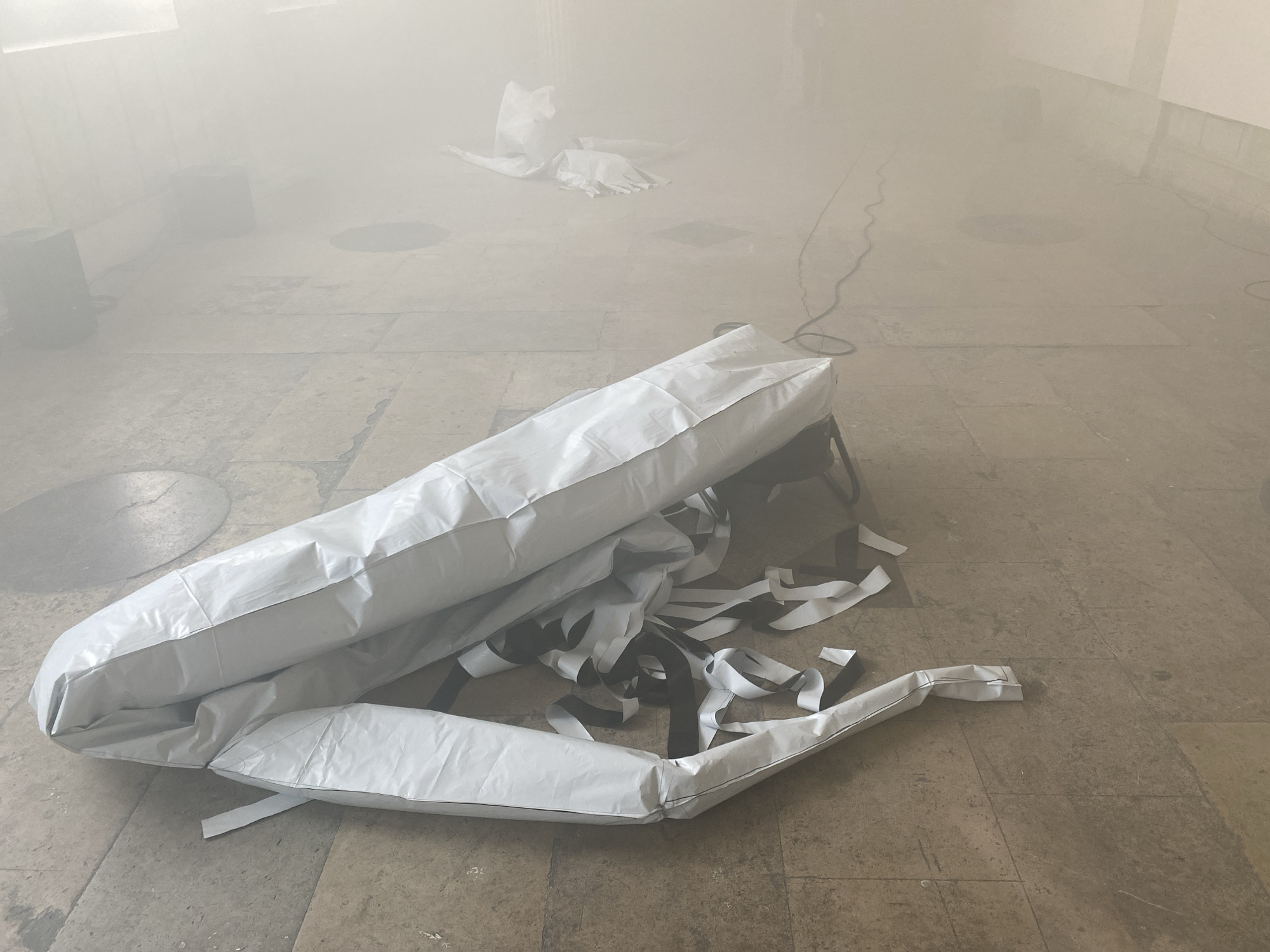 The image depicts an interior space filled with haze, obscuring the background. In the foreground, a deflated, white, pillow-like object rests on a light-colored, tiled floor. Black and white ribbon-like strips are scattered around the object. A second similar object can be seen in the background, albeit less clearly due to the haze. Electrical cabling also lies on the floor. The location is unknown.