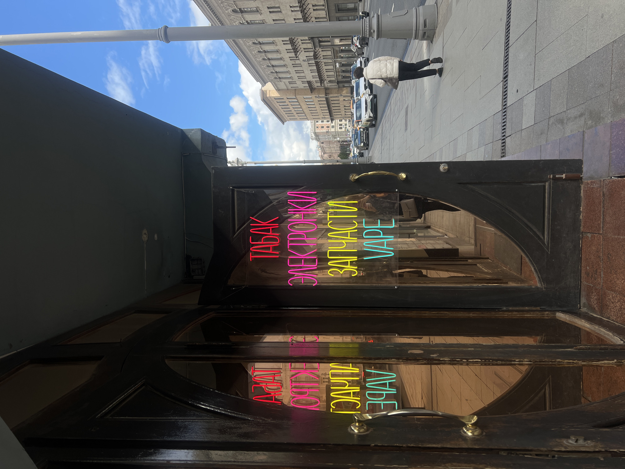 The image shows a street scene in Moscow, Russia. A shop door with a neon sign in Russian stating "Tobacco, Electronics, Spare Parts, Vape" is visible. The sign is in vertical text with different colors: red, magenta, yellow, and cyan. The door is dark, possibly made of wood with glass panels. Reflected in the glass are buildings across the street and a person walking on the sidewalk. The street is lined with parked cars and the sky is partly cloudy. The scene appears to be captured during daylight hours. The photo is taken from a low angle and is rotated counterclockwise.