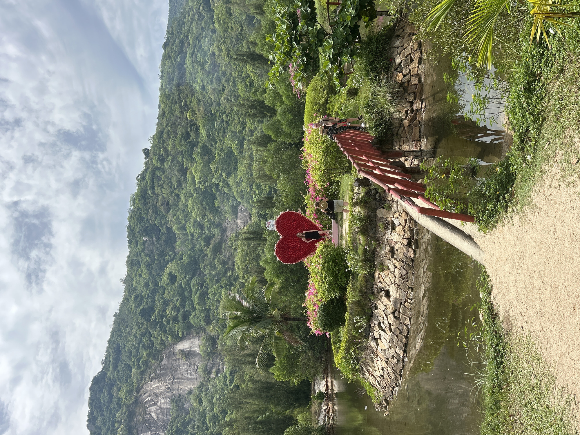 This is a landscape photograph taken at the Thien Mu Pagoda in Hoa Thang, Vietnam. The image features a small, man-made island in a pond, which is decorated with a large, red, heart-shaped sculpture made of flowers. Two people are visible on the island, one standing near the heart sculpture and another walking across a red bridge that connects the island to the mainland. The island is surrounded by a stone wall and lush green vegetation. In the background, a steep, forested mountain rises, with a rocky cliff face on its left side. The sky is cloudy and overcast. The foreground shows a sandy path leading towards the bridge and the island.