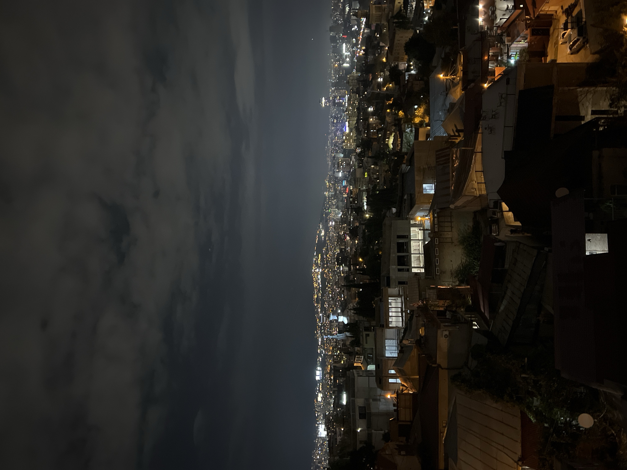 This image captures a nighttime view of Tbilisi, Georgia, from a vantage point overlooking the city. The scene is dominated by the glow of city lights stretching across a sprawling urban landscape under a cloudy sky. In the foreground, residential buildings with various roof styles, some appearing to be older and others more modern with large windows, are visible. The rooftops are a mix of metal and tile, with some showing signs of age.
Further into the distance, the city unfolds with a dense concentration of buildings, their windows illuminated, creating a tapestry of light. A prominent church spire with a faint light emanating from it stands out among the structures, suggesting a religious building within the cityscape. The urban sprawl extends towards the horizon, where the lights become more diffused and blend with the dimly lit sky.
The overall impression is one of a vibrant city at rest for the night, with the artificial illumination highlighting its extensive reach and the topography of the hilly terrain. The presence of some parked vehicles is discernible in the lower right portion of the image, adding a subtle touch of daily life to the nocturnal panorama. The atmospheric conditions, characterized by clouds, contribute to a soft diffusion of light, giving the scene a somewhat subdued yet magical quality.