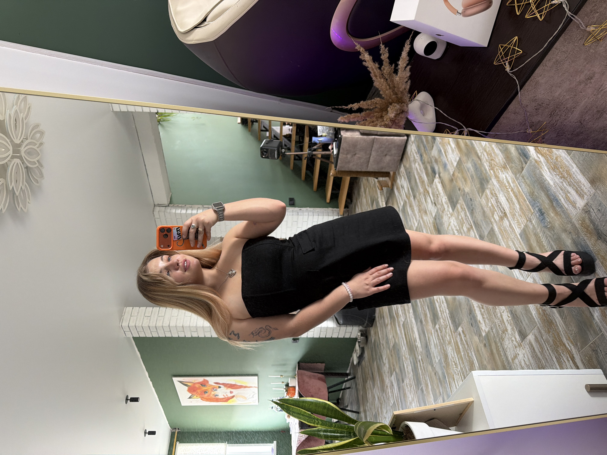 A woman is taking a selfie in front of a mirror. She is wearing a black dress and black sandals. The background shows a room with green and white walls. There is a painting of a fox on the wall. There is a plant in a white pot. The location is Большая Шильна, Russia.