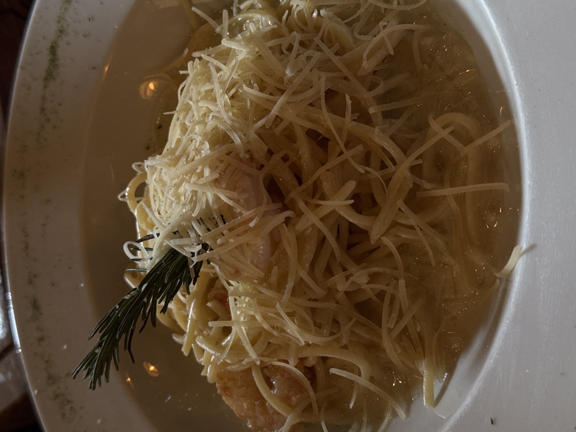 A close-up overhead shot depicts a plate of pasta, generously topped with shredded cheese. A sprig of rosemary adorns the dish. The plate's surface appears to have some scattered green specks.
The location is plausibly Puebla, Mexico, but it cannot be confirmed from the visual information available.