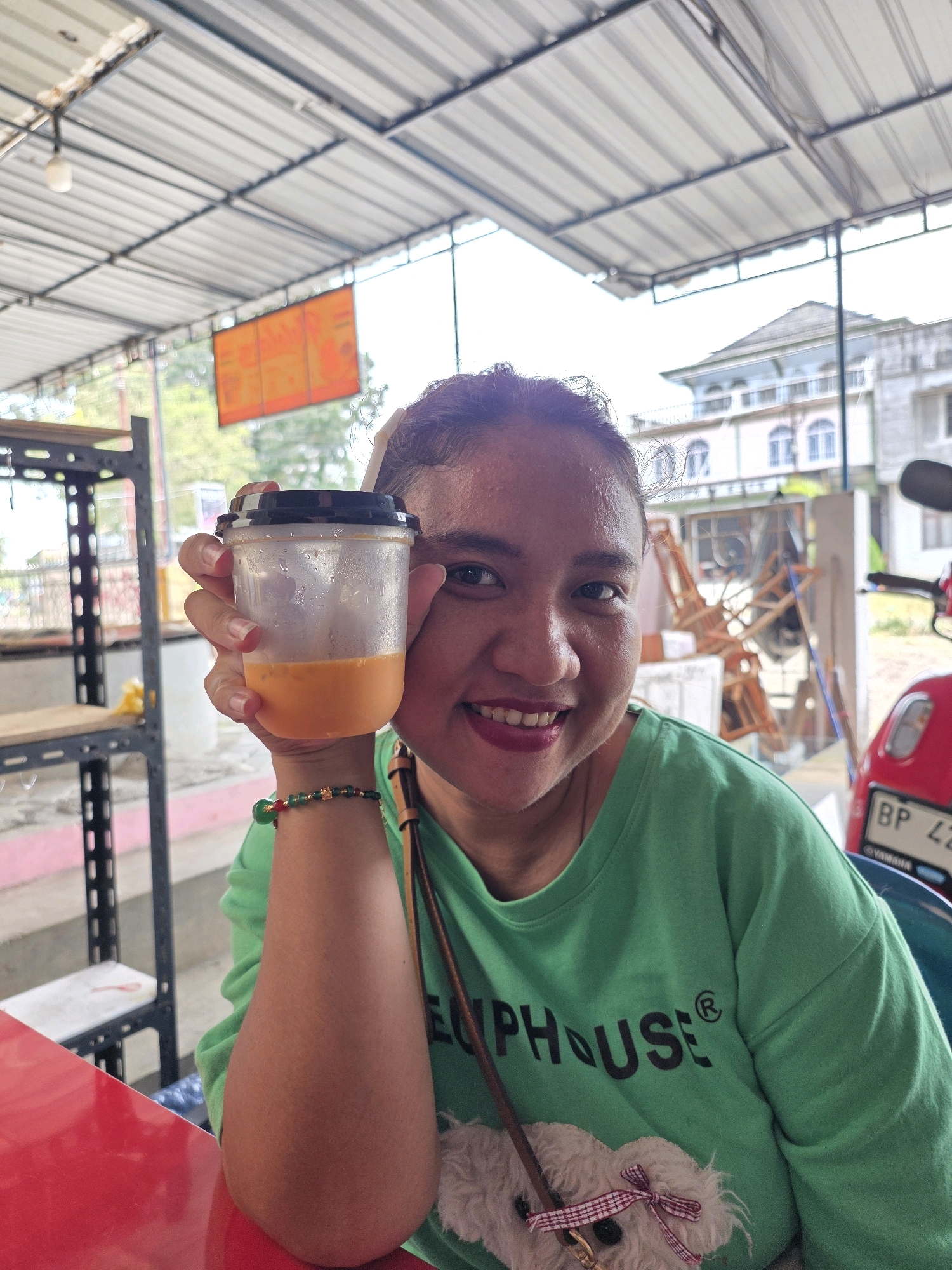 A young woman is holding a plastic cup with an orange beverage to the camera. She is wearing a green t-shirt and has a beaded bracelet on her left wrist. In the background, there is a building with arched windows, a red motorcycle, and a metal shelf. The scene appears to be outdoors, possibly at a roadside cafe or market, with a corrugated metal roof overhead. The location is identified as Tanjung Pinang, Indonesia.