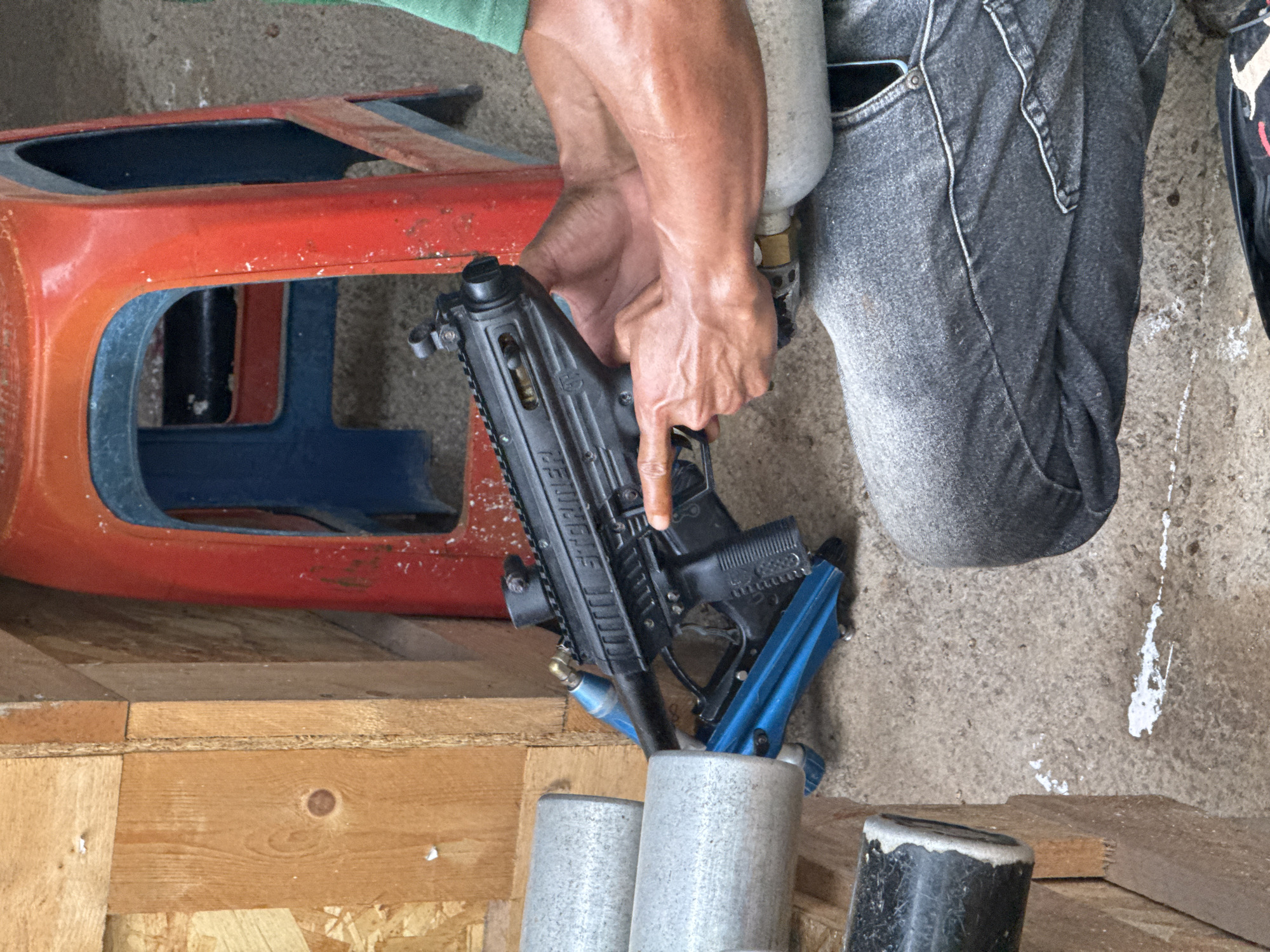 A person is holding a black Tippmann Stormer Elite paintball marker with a blue stock. The marker has an air tank attached and is partially inserted into a collection of cylindrical metal objects. A red and blue object rests on a wooden surface in the background. The person is wearing faded gray jeans and a green shirt. The location appears to be an indoor space, potentially a workshop or garage, with a concrete floor. There is no visible indication that the image was taken in Kuta Selatan, Indonesia.