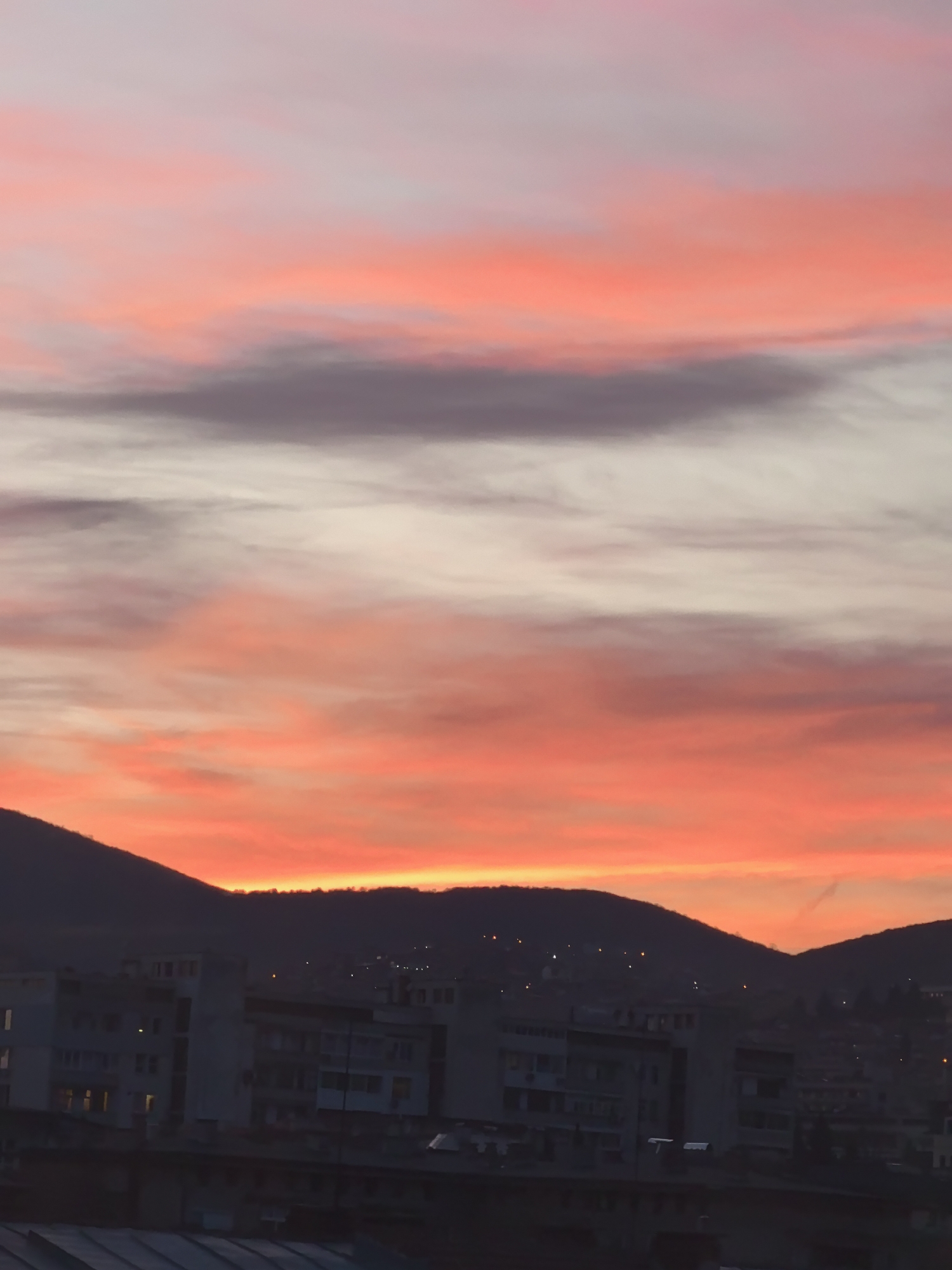 The image shows a vivid sunset sky with hues of red, pink, and gray over the city of Stara Zagora, Bulgaria. In the foreground, there are silhouetted buildings, and slightly behind them are dark mountain ridges. A few lights are visible on the mountains, likely from houses or other structures. The sky takes up most of the frame, exhibiting a dramatic gradient of colors. The buildings appear to be residential or commercial structures. The overall impression is one of a cityscape viewed during the twilight.