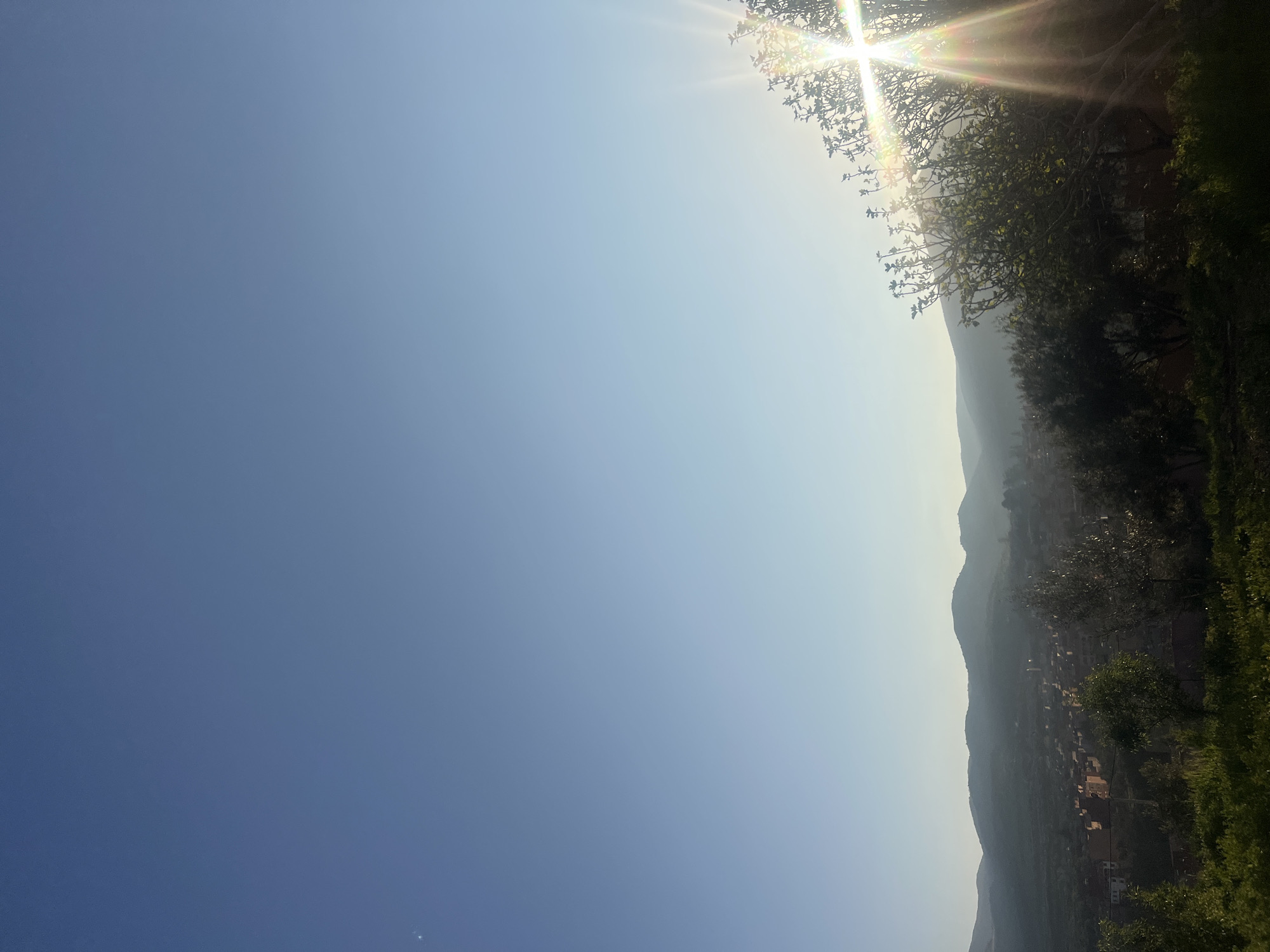 The image captures a broad landscape view of Ouaouizert, Morocco, under a bright, sunlit sky. The foreground is dominated by the sky, transitioning from a deep blue on the left to a lighter, hazy blue towards the horizon. The sun is visible in the upper right, obscured slightly by the branches of a tree. The landscape consists of a mountain range. Buildings within the village are visible among the landscape. Lush green vegetation and trees frame the lower right part of the image.