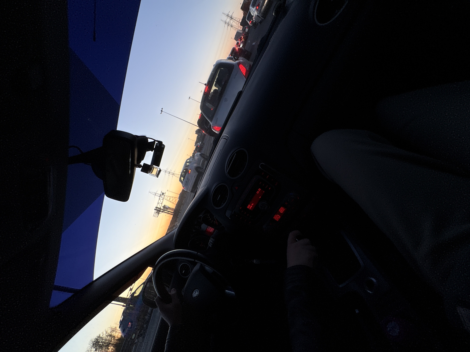 From inside a car, a driver is stuck in traffic at dusk in Kudrovo, Russia. The car's dashboard is visible with red electronic displays. Numerous cars are ahead, stopped or moving slowly on the road. Power lines and city infrastructure are visible through the windshield. The sky is light with the sun setting. The driver is holding the steering wheel and another hand is near the gear shift.