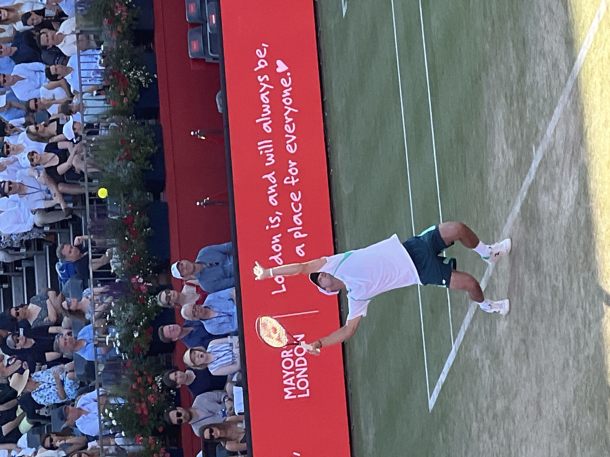A male tennis player in a white shirt and dark shorts is captured mid-serve on a green tennis court. He is positioned on the right side of the frame, with his left arm extended upwards, holding a tennis racket behind his head as he prepares to strike a ball. His body is slightly contorted, showcasing the athleticism of the moment. The court has white lines marking its boundaries. In the background, a red wall displays white lettering that reads "MAYOR LONDON" on the left and "London is, and will always be, a place for everyone." followed by a pink heart symbol on the right. Behind the wall, spectators are seated in tiered seating, many of whom are blurred or out of focus. The overall setting appears to be an outdoor tennis match at a stadium, likely in London given the prominent signage.