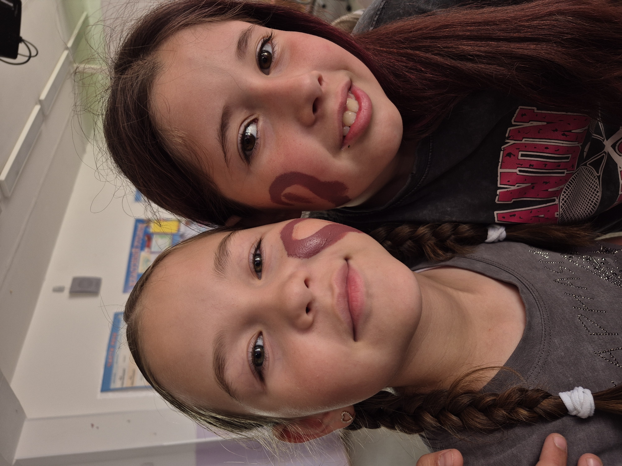 The image shows two young girls, likely friends or siblings, with face paint. They are positioned close together, with one girl's head resting on the other's shoulder. The girl in the foreground has brown hair in braids and a grey t-shirt. She has a heart-shaped earring in her left ear. The girl behind her also has brown hair and is wearing a dark t-shirt with red and white lettering that appears to spell "VINTON" in a distressed font, along with a graphic of a tennis racket. Both girls have circular, dark red face paint designs on their cheeks.
The setting appears to be indoors, possibly a school or a community center, suggested by the white walls and posters on the left side of the image. The lighting is bright, indicating it is daytime. The overall mood is playful and cheerful, with the girls smiling and looking at the camera. The image is an upward-angle shot, taken very close to the subjects.