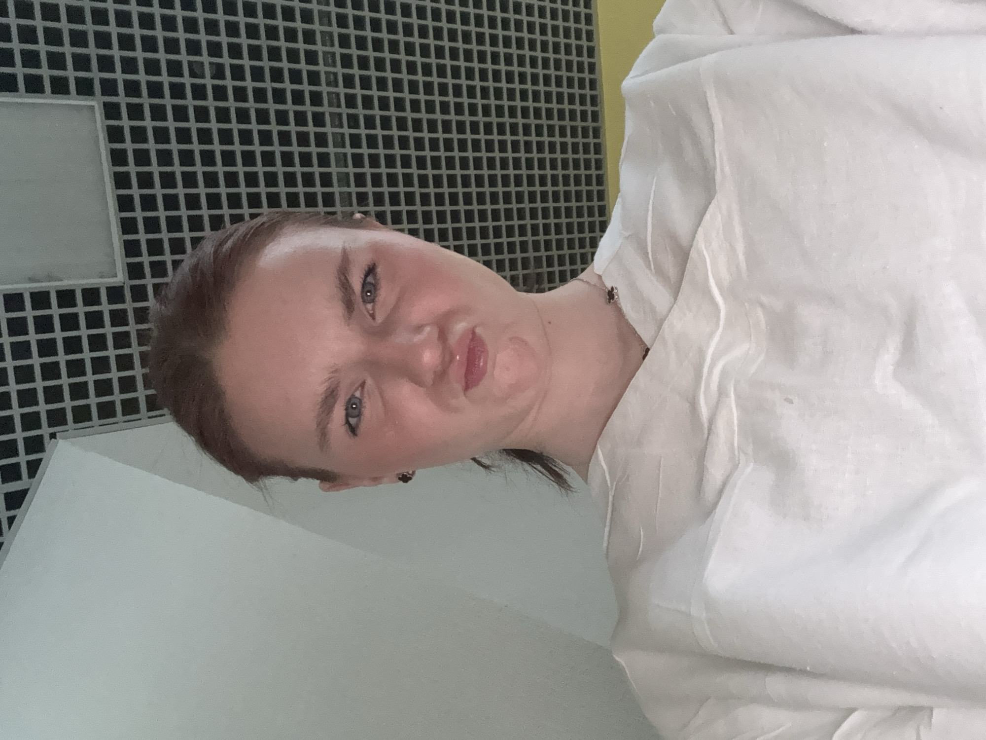 A young woman is pictured indoors, possibly in a bathroom, given the tiled wall in the background. She is wearing a white top and has her hair pulled back. She's making a pouty face directly at the camera. The location is unknown, and there is no explicit evidence to suggest it is in Saint Petersburg, Russia.
