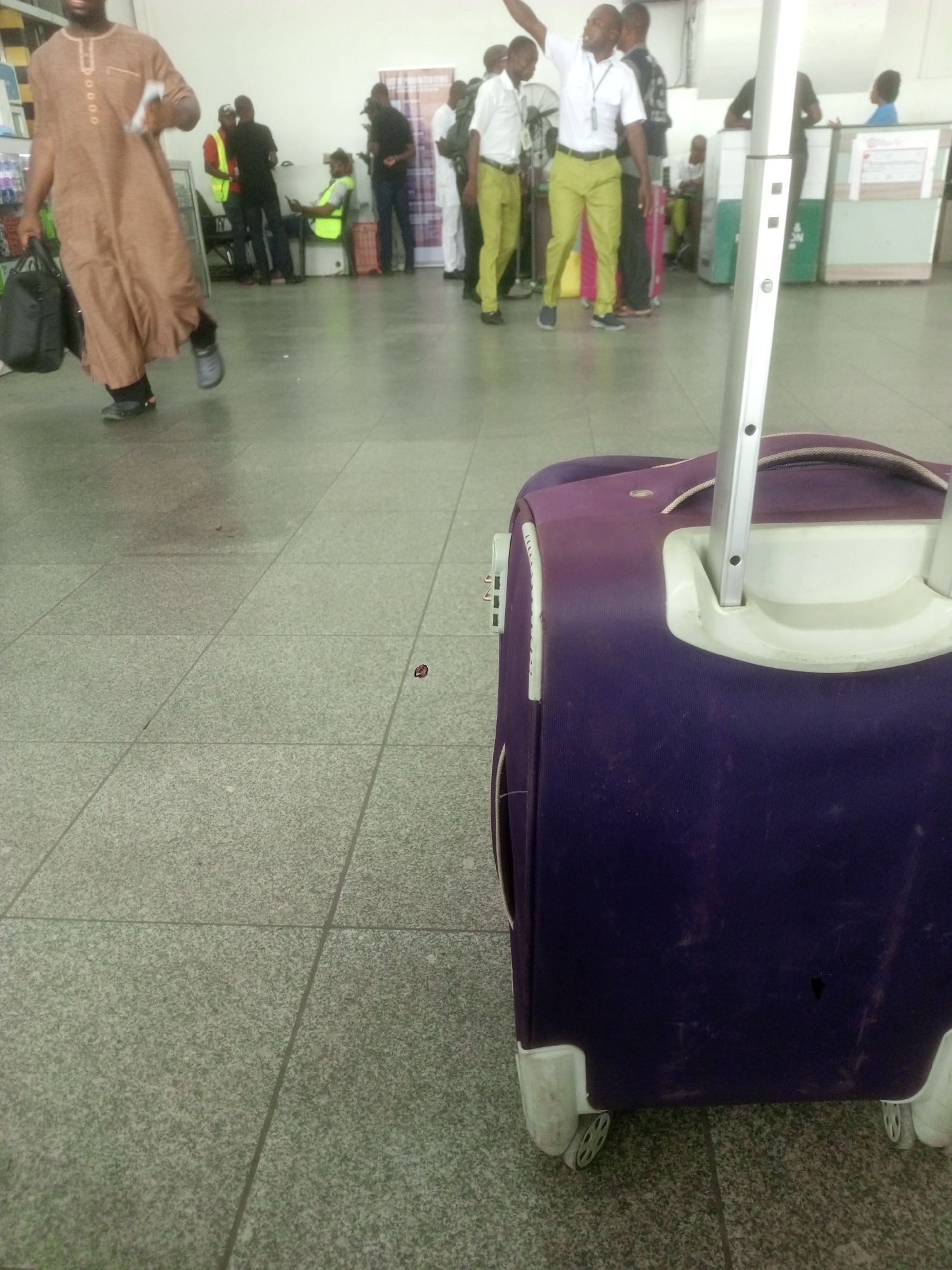 The image depicts an airport scene in Abuja, Nigeria. In the foreground is a purple rolling suitcase with its handle extended. The floor is tiled. In the background, a group of people are visible. One man in a brown robe carries a black bag and walks towards the left. Another man in a white shirt and chartreuse pants has his arm raised. There are several other individuals in business casual attire standing or sitting near what appears to be a counter or service area.
