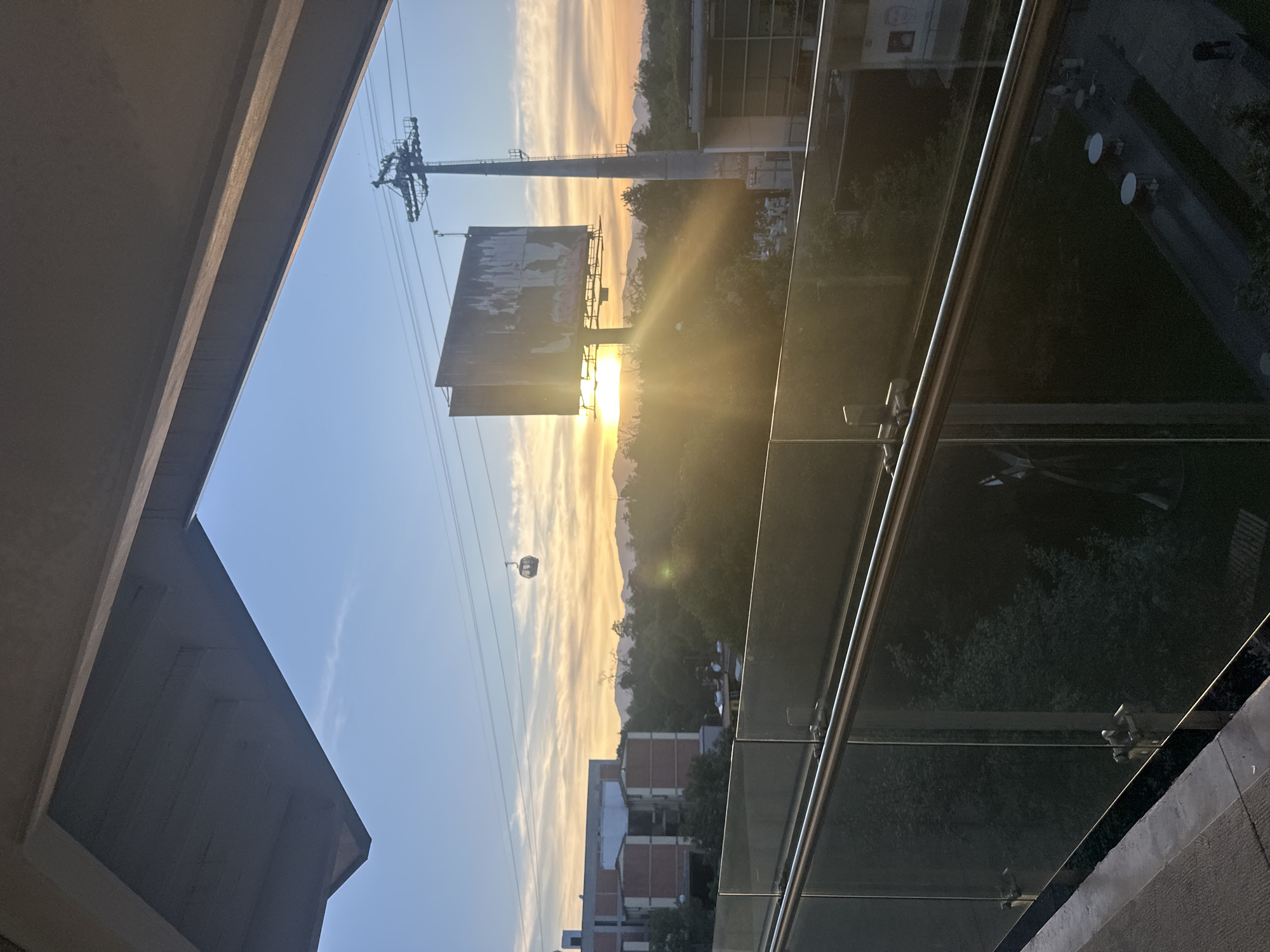 The image shows a sunset view in Mexico City, Mexico, including an elevated cable car system (Cablebús). A large billboard and cable car tower are silhouetted against the setting sun. A cable car is suspended along the cable. The foreground includes elements of a building structure, possibly a balcony or window frame, with glass railings reflecting the surrounding environment. In the distance, buildings and trees are visible.