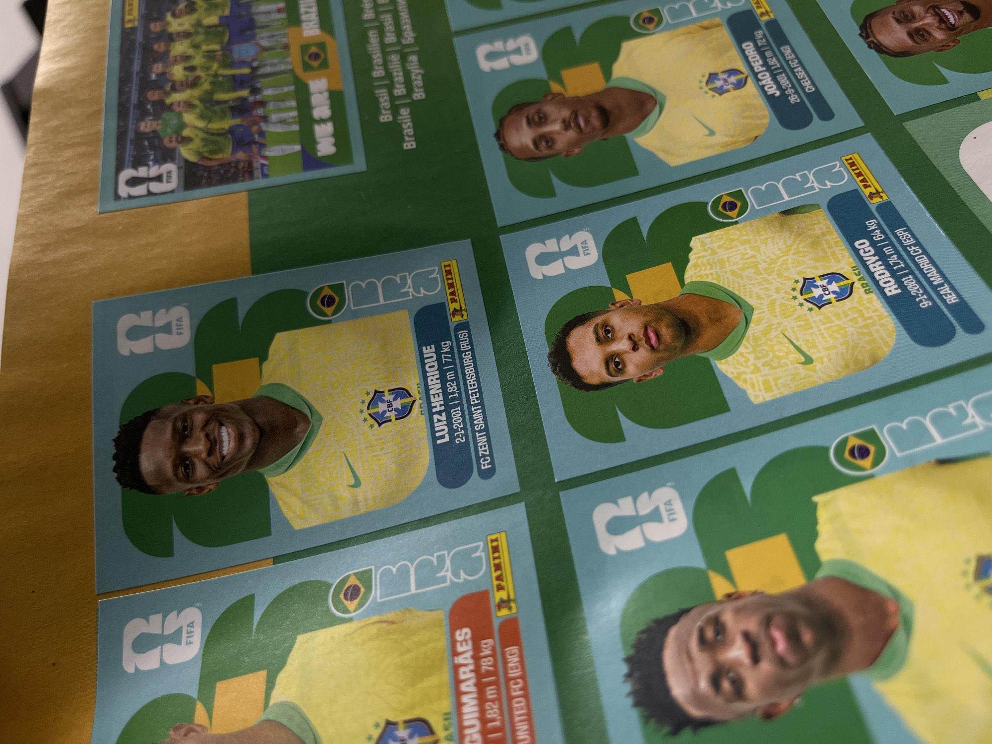 The image shows a collection of FIFA Panini stickers featuring Brazilian football players. From left to right, the stickers show Bruno Guimarães, Luiz Henrique, the Brazilian national team, Rodrygo, and João Pedro. Each sticker includes a photo of the player in the Brazilian national team jersey, along with their name, date of birth, height, weight, and club information. The stickers are arranged on a surface that appears to be either a desk or table with a slightly reflective surface.