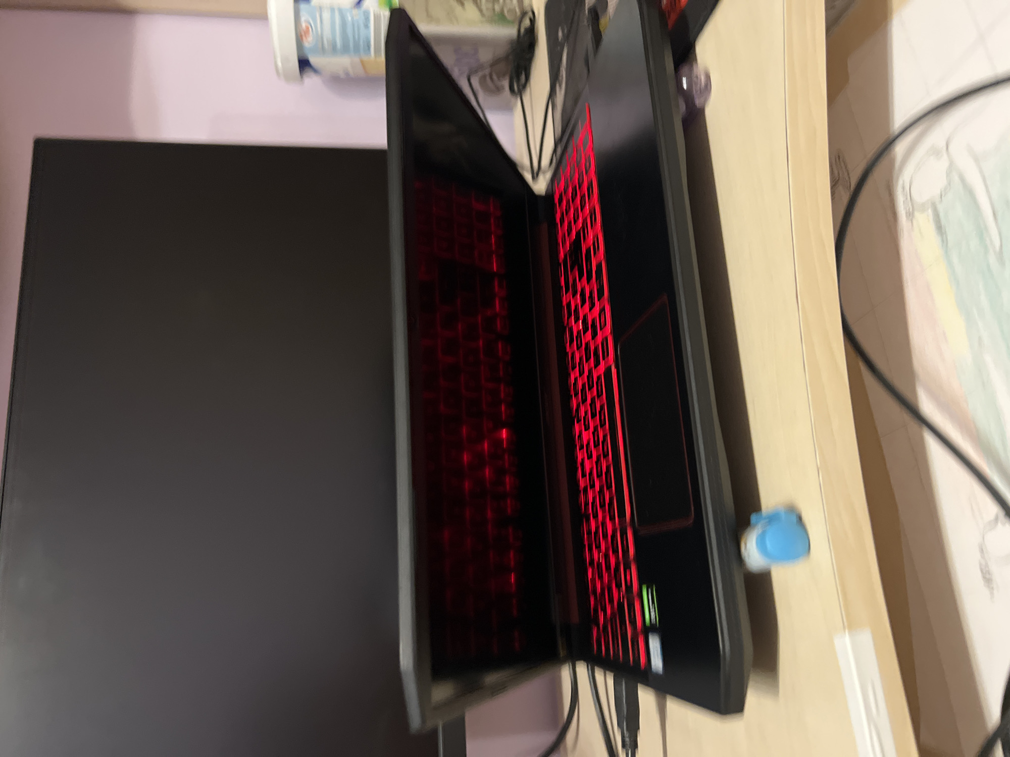 The image displays a personal computer setup, rotated 90 degrees clockwise, showing a desk with a monitor and an open laptop. When viewed with the desk horizontally oriented, the scene reveals:
On the left side of the desk is a large, black, flat-screen computer monitor. Its screen appears dark or powered off. To its right, positioned centrally on the desk, is an open laptop. The laptop features a prominent red backlit keyboard, with individual keys illuminated in bright red, suggesting it may be a gaming laptop. The laptop's screen is also dark or off.
The desk surface is a light-colored, possibly wooden or laminate material. Numerous black cables are visible, originating from the laptop and potentially the monitor, trailing towards the back of the desk. A small, blue and white USB dongle or device is plugged into the right side of the laptop (when viewed from the front).
In the background, behind the desk setup, is a solid light lavender or pale purple wall. On a shelf or ledge against this wall, a white container with a blue cap or lid is partially visible. Faint, illegible text is present on the container. Further to the right on the desk, a piece of paper with a faint drawing or pattern, showing hints of green and yellow, is visible, possibly taped to the wall or lying flat.
The overall lighting is ambient, with the red keyboard backlighting providing a distinct internal glow. No specific time of day can be determined, but the indoor setting is clear. The visible text on the laptop keyboard includes standard keys like "Fn", "Shift", "Ctrl", "Alt", and the "Windows key".