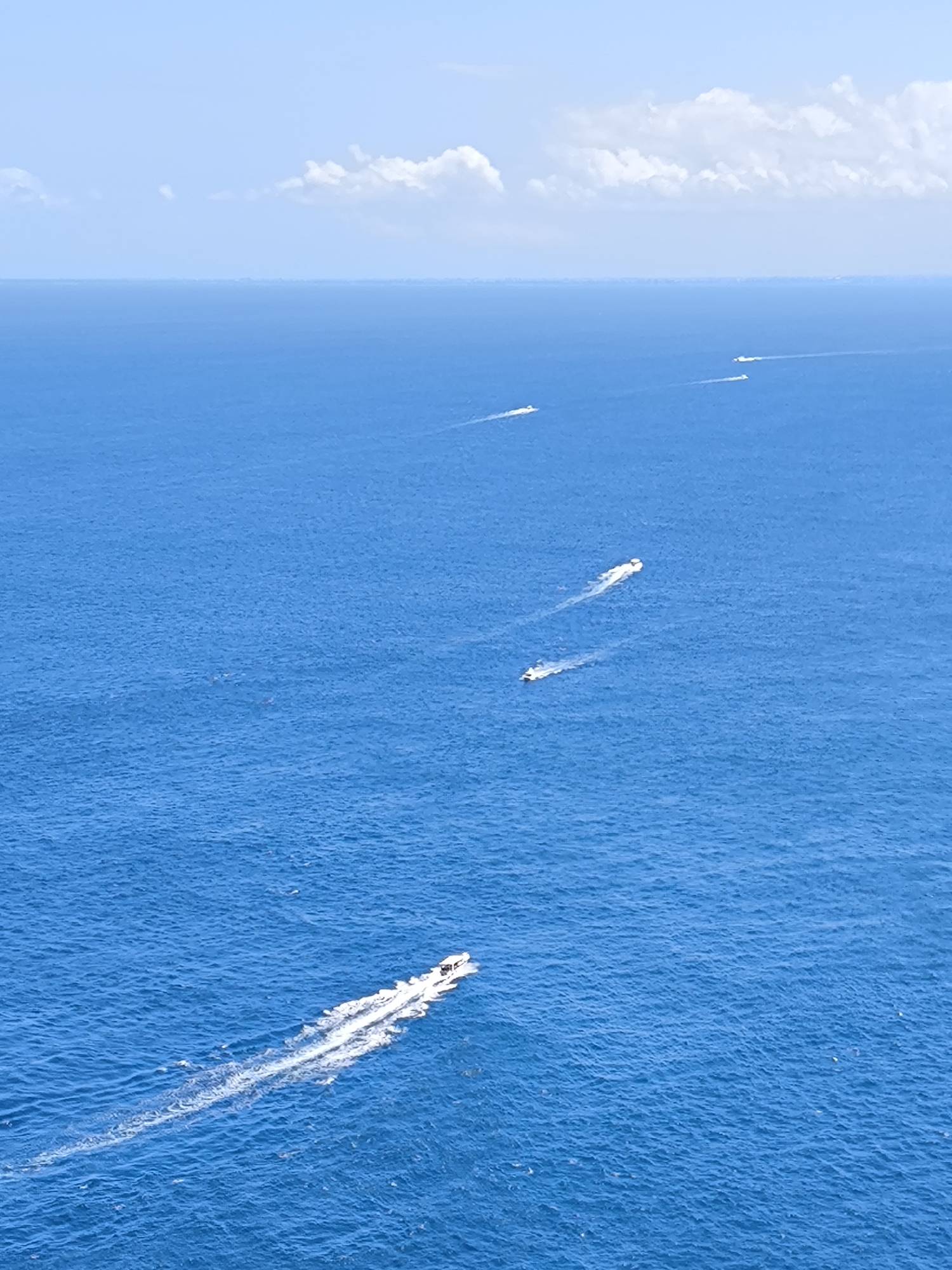 A high-angle outdoor shot captures a vast expanse of deep blue ocean under a clear, bright sky with scattered white clouds. The horizon line, where the sea meets the sky, is visible in the upper portion of the frame.
Several small motorboats are visible across the water, each leaving a distinct white wake behind it, indicating they are moving forward. The closest boat is positioned in the lower-right foreground, angled towards the upper left, displaying a prominent, foamy white trail in its wake. Further into the distance, a line of four more boats can be seen, progressively smaller due to perspective. These boats also generate visible wakes.
The water surface shows subtle ripples and texture, reflecting the bright daylight. The overall impression is one of calm yet active movement on the open sea. The time of day appears to be bright daytime, likely midday or early afternoon, given the strong illumination and high sky. The weather is fair and sunny. The scene is set in the open waters near Nusa Penida, Indonesia. There is no visible text in the image.