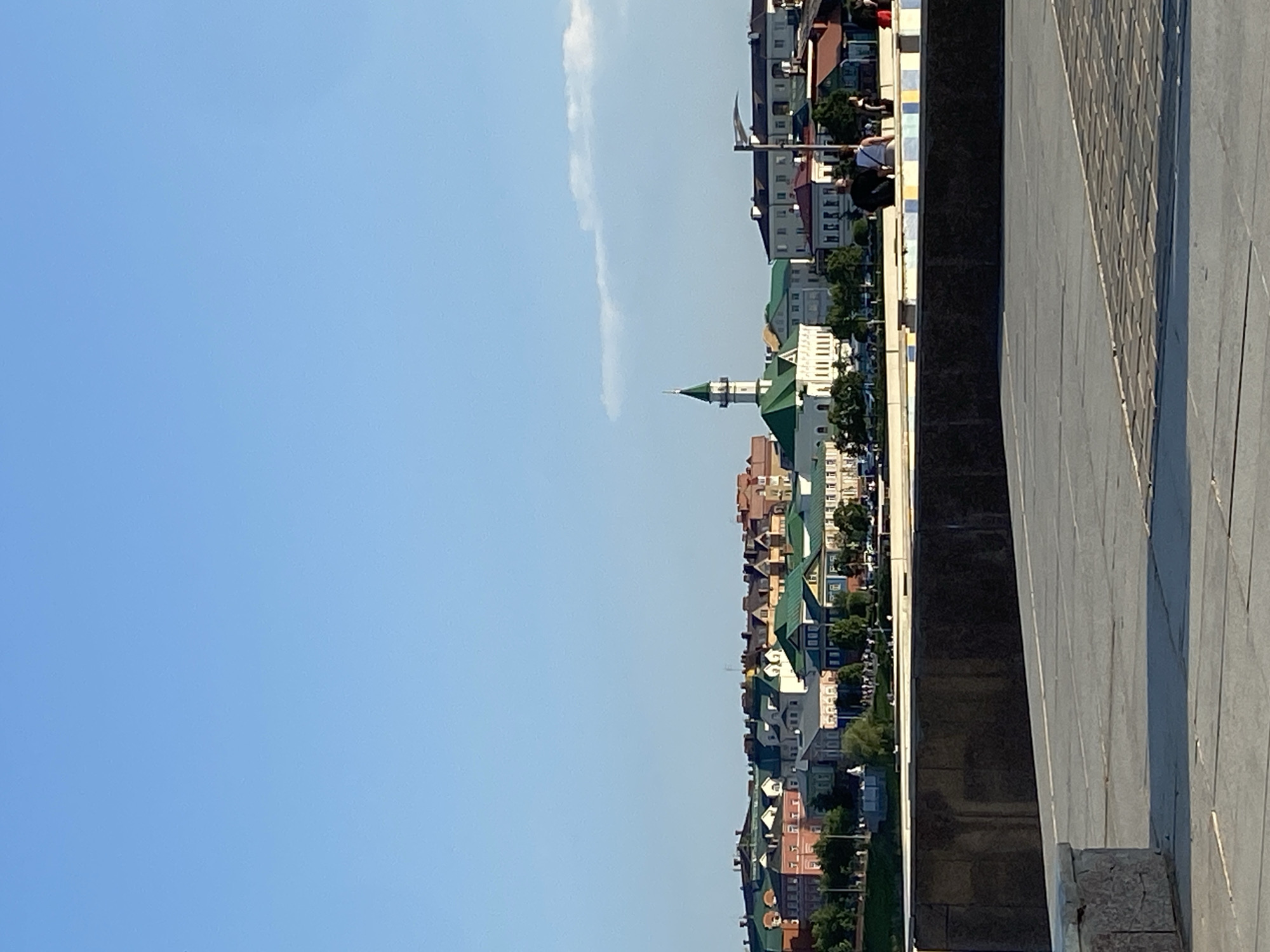 The image shows a waterfront cityscape in Kazan, Russia, on a clear, sunny day. A row of colorful buildings, many with green roofs, stretches along the waterfront. A white mosque with a green spire stands out among the buildings. Pedestrians sit and stand at the water's edge, alongside decorative edging and pavement. The sky is a clear, light blue with a thin, vertical cloud. The image is taken from a slightly elevated perspective.