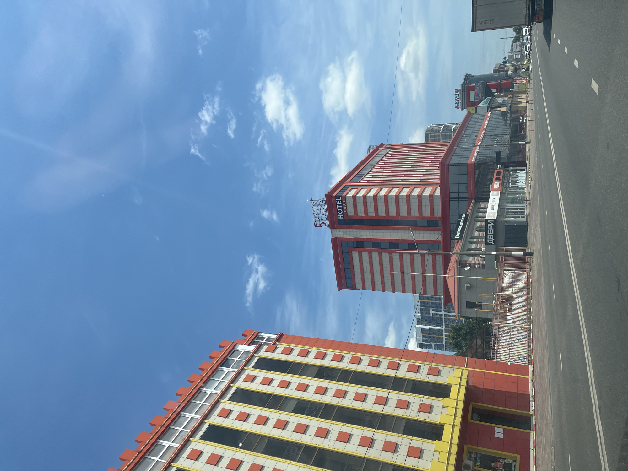 This image depicts a street view in the city of Krasnodar, Russia, on a clear, sunny day with a bright blue sky and scattered clouds. The foreground shows a multi-lane asphalt road with white dashed lines marking the lanes. On the right side of the road, a white truck with a trailer is visible, partially obscuring other vehicles in the distance, including several white cars.
The buildings lining the street are the main subjects of the image. On the left, a strikingly colorful building with a bright red exterior and yellow accents is prominent. It features multiple stories with rows of windows, interspersed with square red decorative elements. The building has a decorative red crenellated top.
Further down the street, a taller, modern building with red and white horizontal stripes dominates the skyline. This building is identified by a sign at the top as "FORUM PLAZA" and also displays "HOTEL" with four stars, indicating it is a hotel. Below the hotel, there is a commercial area with storefronts. One sign reads "ДВЕРИ" (Doors). Another sign visible on a building further down the street reads "МАЛЫШ" (Baby/Kid).
The architecture suggests a mix of older and newer commercial and residential developments. The overall impression is a bustling urban environment under a clear sky.
