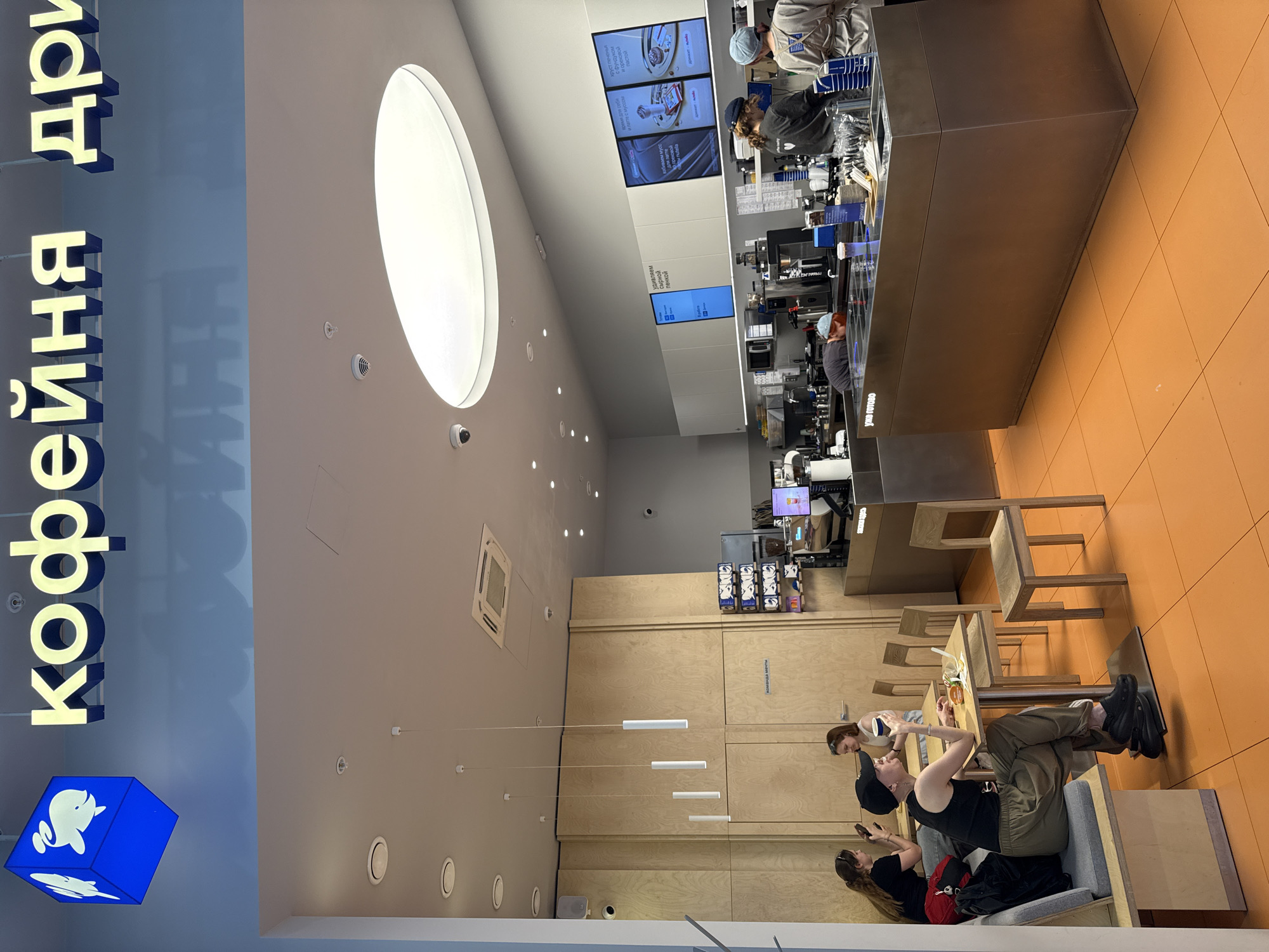 Here is a detailed description of the image:
**Scene:** The image shows the interior of a coffee shop, likely in Moscow, Russia, based on the Cyrillic writing ("кофейня дри") on the sign. The view is from a slightly elevated and angled perspective, looking towards the ordering counter.
**Environment:**
* The interior is modern with a neutral color palette and minimalist design.
* The ceiling is high with recessed lighting and a large oval skylight.
* One side of the coffee shop has light wooden paneling.
* The floor in some areas is a bright orange/yellow tile.
* Pendant lights with long, slender cylindrical shades hang over the seating area.
**People:**
* There are a few people in the coffee shop.
* Two baristas are visible behind the counter, preparing drinks.
* Three customers are sitting in a lounge area, possibly looking at phones.
**Objects:**
* There is a counter with multiple screens displaying food and drink items in Russian.
* A display case with coffee equipment is visible behind the counter.
* A few wooden chairs and tables are positioned in the seating area.
* There are various personal belongings on the chairs, such as bags.
**Actions/Interactions:**
* The baristas are working behind the counter.
* The customers are sitting and relaxing, potentially using their phones or conversing.
**Specific Details:**
* One of the customers is lying down on a bench.
* The menu displays on the screen showcase coffee drinks.
* A blue sign featuring a logo is displayed above the shop.