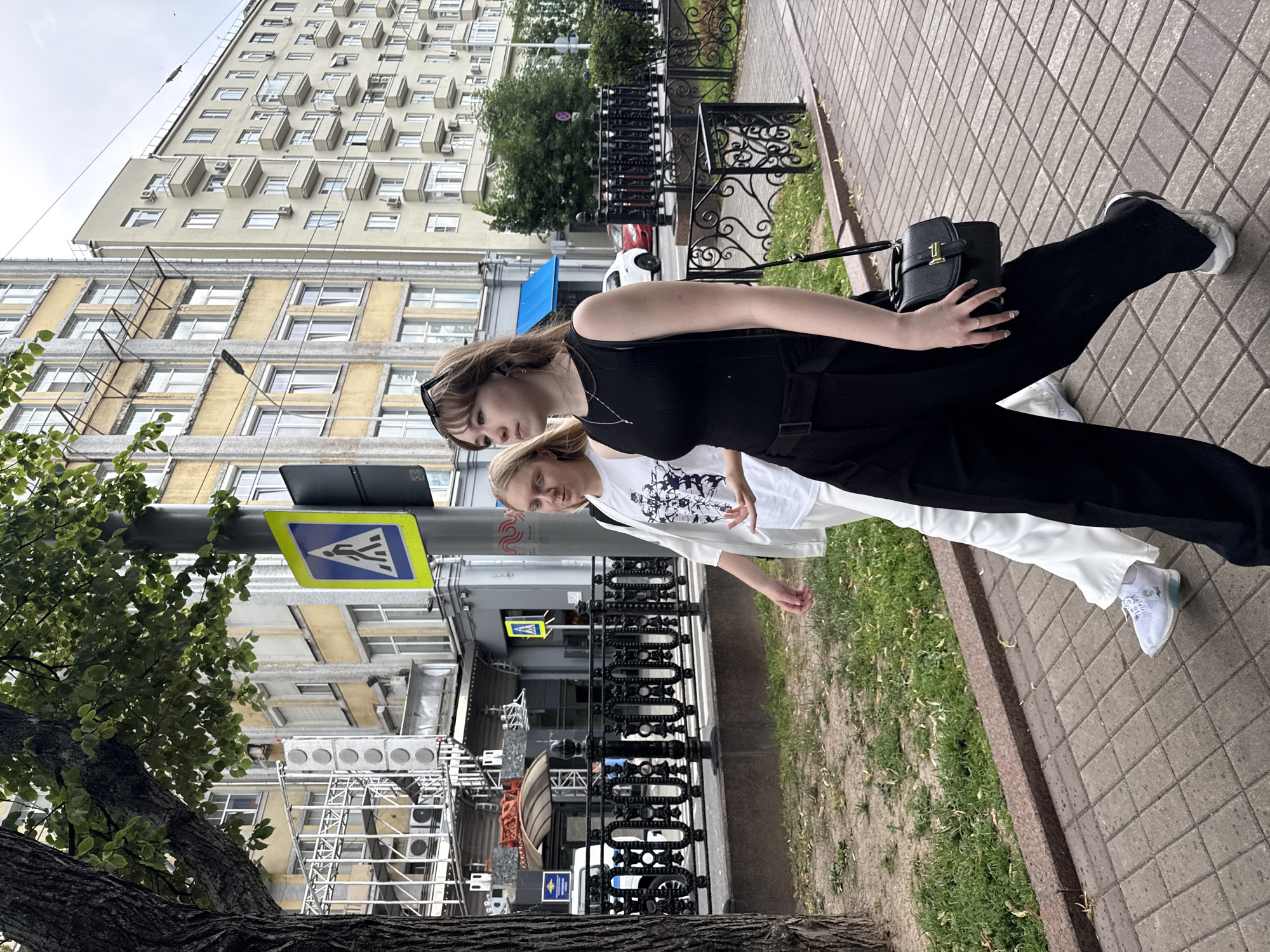 The image depicts two young women walking along a sidewalk in Moscow, Russia. The woman in the foreground is wearing a black sleeveless top and black pants, and her hair is light brown. She is looking down and to the side, with sunglasses perched on her head. She is carrying a small black handbag. The woman behind her is wearing a white t-shirt with a graphic print and white pants.
They are walking on a sidewalk made of light brown paving stones, bordered by a patch of green grass and a stone curb. In the background, there is a tall, light-colored building with many windows and balconies, typical of Soviet-era architecture. A pedestrian crossing sign with a blue background and a white silhouette of a person walking is visible on a pole to the left of the women. Further in the background, there are more buildings and trees. The scene appears to be set during the daytime, with overcast skies, as indicated by the diffuse lighting. The overall mood seems casual and relaxed, suggesting they are out for a stroll. No discernible text is visible beyond the pedestrian crossing sign.