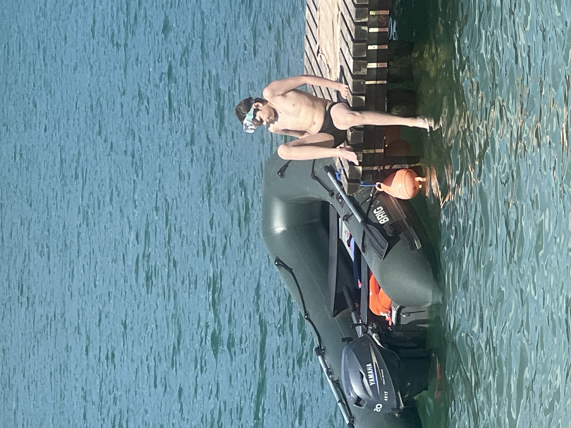 A young person in black swim trunks and a snorkeling mask is crouching on the edge of a wooden dock. The dock is made of weathered planks and extends over calm, blue-green water. Beside the dock, a dark green inflatable boat with a Yamaha outboard motor is partially submerged. An orange buoy is tied to the boat. The water is slightly rippled, reflecting the sunlight, suggesting it is a bright day. The scene appears to be a recreational setting, possibly a lake or a river, with the person preparing to enter the water for snorkeling or swimming. The overall mood is relaxed and summery. The visible text on the boat's motor reads "YAMAHA" and "20 4815".