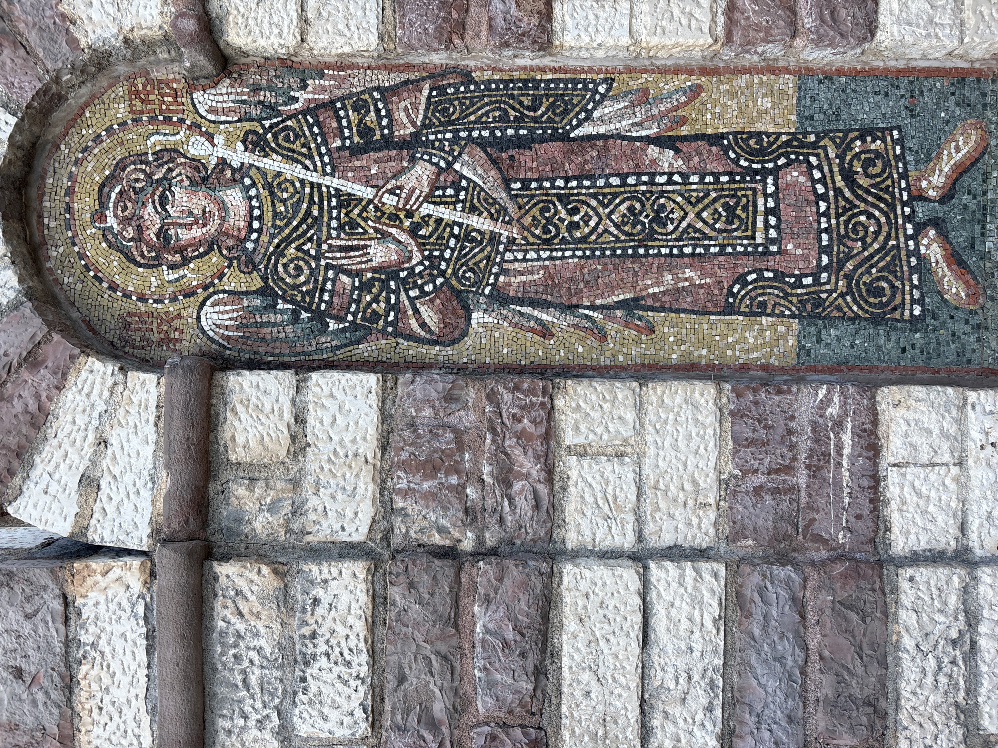 The image depicts a mosaic of an angelic figure on the exterior wall of a building in Budva, Montenegro. The mosaic is set within an arched recess in a stone wall constructed with alternating bands of lighter and darker, rough-hewn stones. The angelic figure is depicted frontally, wearing a richly patterned robe and holding a staff. Its face is rendered with an expression of serenity. The mosaic uses a variety of small, colored tiles to create the image, with a halo surrounding the figure's head. The surrounding stone wall appears weathered, with some stones exhibiting signs of age and erosion. The overall impression is one of historical and artistic significance, typical of religious or historical architecture found in the region.