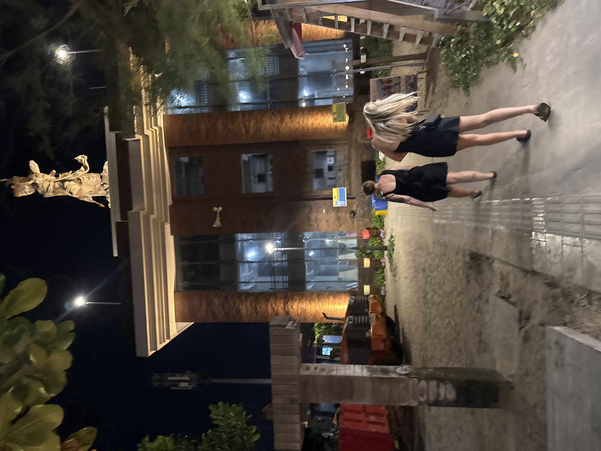The image shows a street scene at night in Kuta, Indonesia. Two women are walking away from the camera on a paved walkway. The woman on the left is wearing a black sleeveless dress and the woman on the right is wearing a black halter dress and has blonde hair. Behind them is a large brick building with illuminated windows and a statue on top. To the left of the building, a large orange construction vehicle is visible. The ground is sandy, suggesting proximity to a beach. Trees and streetlights are also present in the scene.