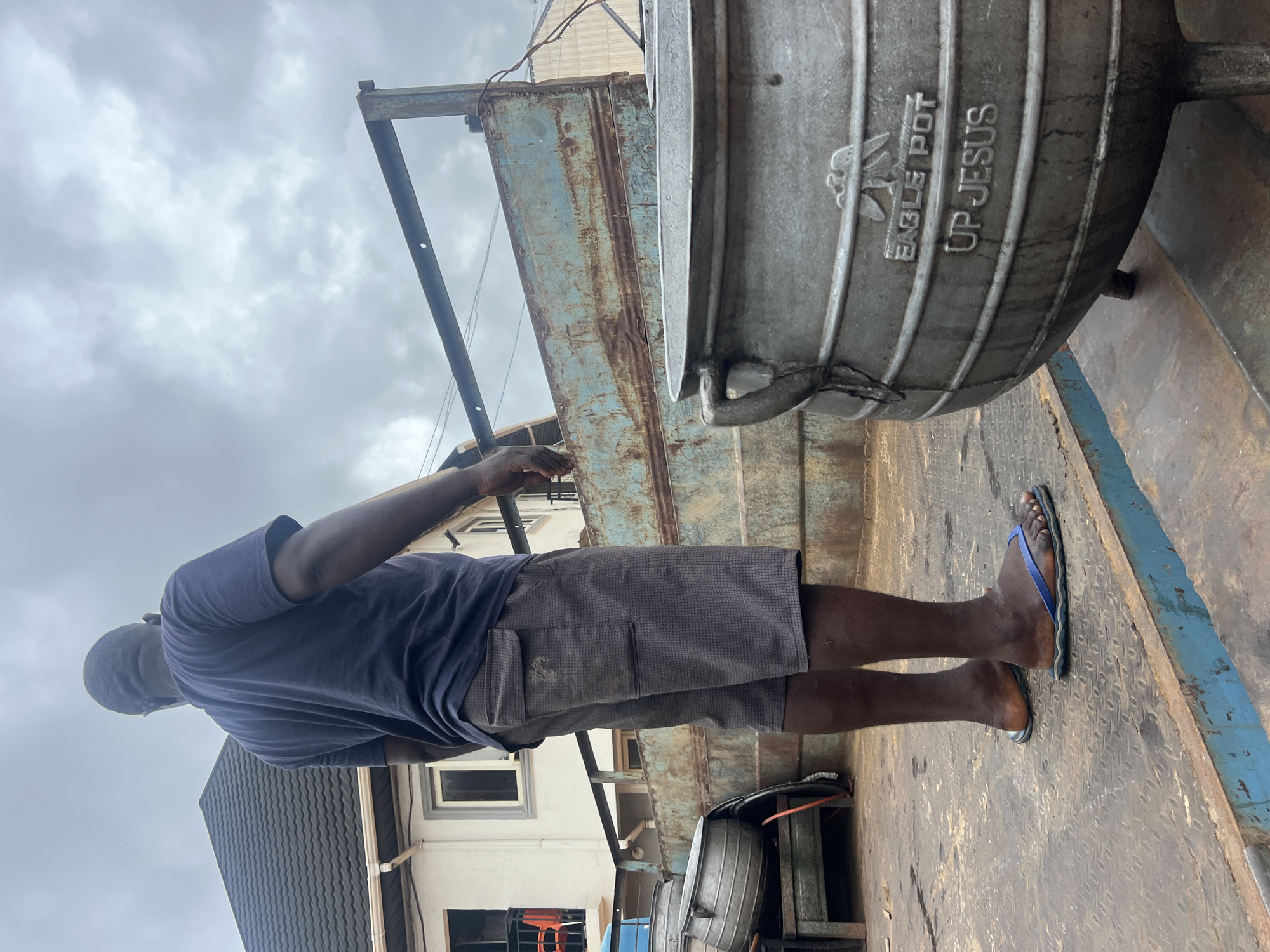 The image depicts an outdoor scene, likely in Onitsha, Nigeria, featuring a man standing next to a large metal pot. The man, with dark skin and short dark hair, is viewed from his left side and slightly from behind. He wears a dark blue t-shirt, dark grey patterned shorts with a cargo pocket, and blue flip-flops.
A prominent feature is a large, cylindrical, metallic pot, possibly made of aluminum, with horizontal ridges and sturdy handles. Embossed on its side are the words "EAGLE POT" above an emblem of an eagle, and "UP JESUS" below. This pot sits on a heavily rusted and weathered blue-grey metal surface, which appears to be a platform or the bed of a truck. Another smaller, dark object, possibly a tire or part of equipment, is partially visible on the ground.
In the background, under an overcast sky filled with grey clouds, are buildings, including a white structure with visible windows and a building with a dark, ridged roof. The overall environment suggests a functional, possibly commercial or industrial, outdoor space during daytime. The man is standing still, observing or waiting; no specific activity is clearly evident.