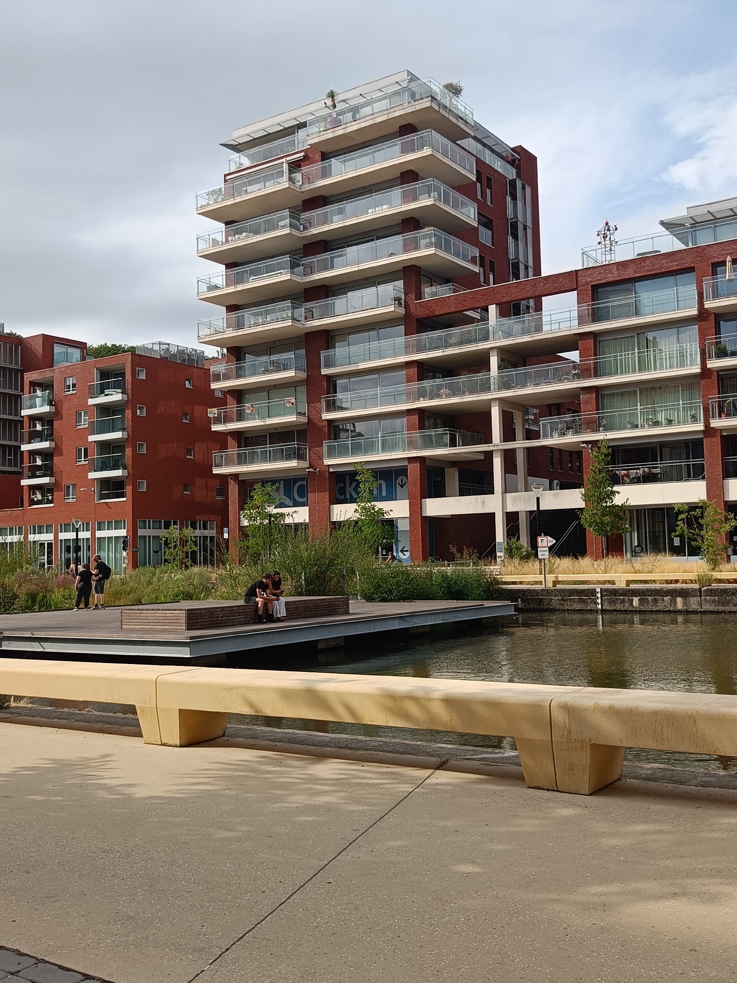 This image depicts a modern urban waterfront scene in Leuven, Belgium, likely near the Vaartkom area, characterized by contemporary red-brick apartment buildings with multiple balconies and large windows. In the foreground, a long, light-colored concrete bench runs parallel to a paved walkway. Beyond the bench, a wooden deck extends over a body of water. On the deck, three individuals are seated on a wooden structure, two women and a man. To the left of the deck, two men are standing and conversing. The buildings exhibit a tiered architectural style, with glass railings on the balconies. Lush greenery and tall grasses surround the waterfront area, contrasting with the man-made structures. The sky is overcast with patches of blue peeking through.