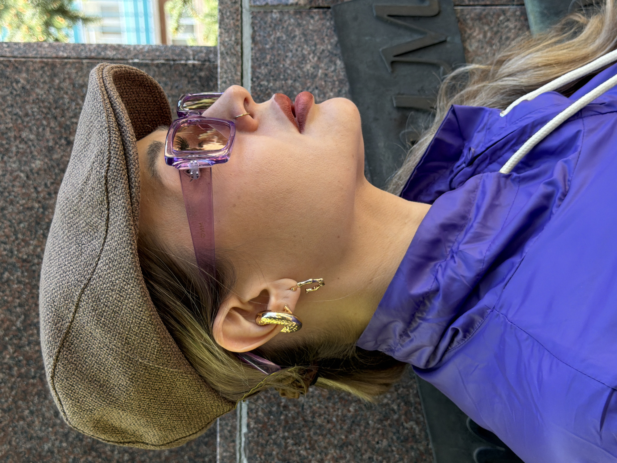 A woman is visible in the image, angled with her head pointing upwards. She wears a brown tweed flat cap, purple-tinted sunglasses, and has a gold nose ring. She also has two gold earrings in her left ear. She is wearing a shiny purple jacket with white drawstrings. The background includes a granite structure and foliage, which suggests an outdoor urban setting.
Based on the style and texture of the surrounding granite structure, along with visual similarities from publicly available images, it is plausible that this image was taken in Krasnoyarsk, Russia, in an area with a monument or statue. The available information suggests the statue is the statue of Andrei Dubensky.