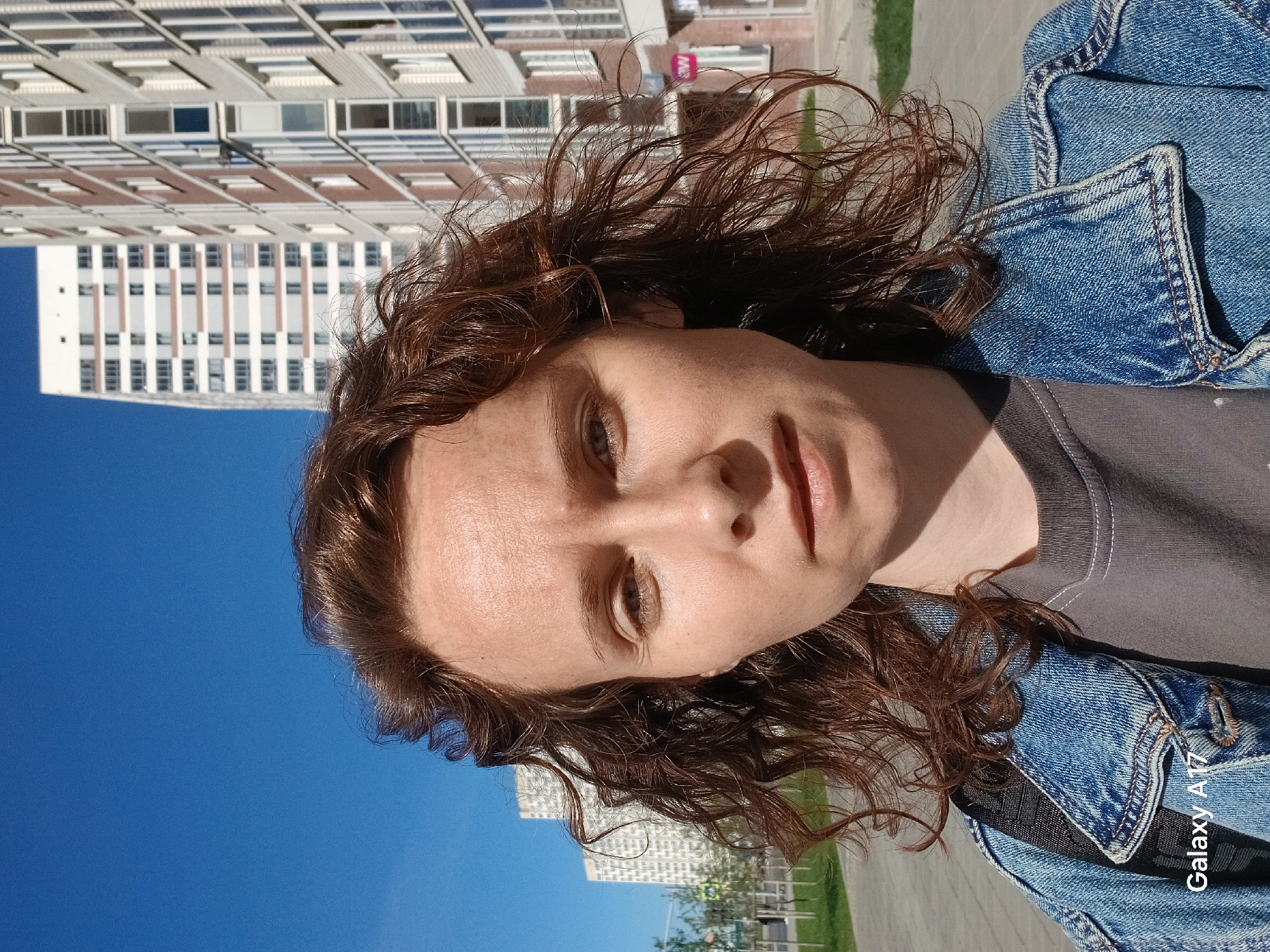 A selfie-style photograph features a light-skinned person with curly, auburn hair wearing a grey t-shirt and a blue denim jacket. The background includes a tall, modern building with a white and brown facade and multiple windows. A clear blue sky is visible. The location appears to be outdoors, likely a street in a city, with some greenery visible in the background. The words "Galaxy A17" are visible in the lower right corner. The city of Yekaterinburg, Russia, cannot be confirmed from the details provided.