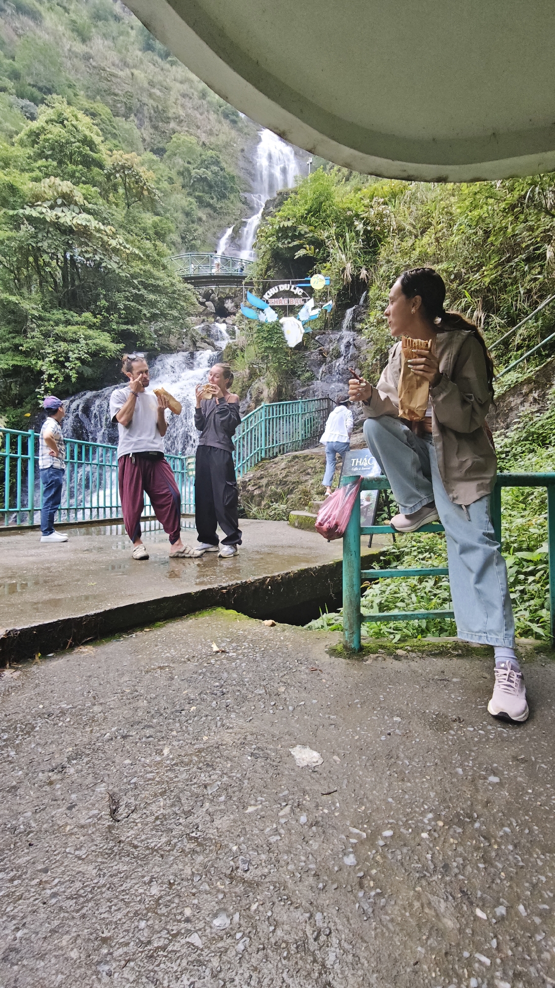 The image captures a casual scene in what appears to be a park or recreational area in Sa Pa Ward, Vietnam. In the background, a cascading waterfall provides a natural backdrop, complemented by lush green foliage and a small footbridge.
In the foreground, a group of people are observed interacting and eating. One woman is sitting on a green metal railing, holding a paper bag and eating, while a couple stands near a green metal fence, also eating. Another person is standing to the left of the couple. The ground is wet, suggesting recent rain. The overall atmosphere is relaxed and informal.