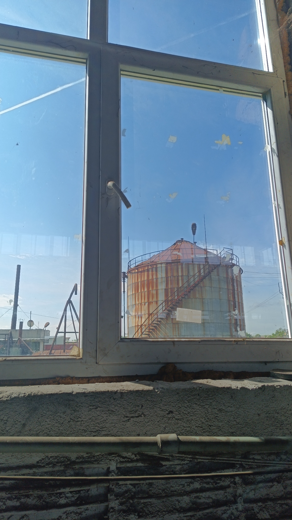 A view through a multi-paned window reveals a large, cylindrical storage tank or silo. The tank exhibits extensive rust, suggesting age and weathering. A staircase winds around the side of the tank. The sky visible through the window is clear and blue. In the left pane of the window, there are various structures and a pole, possibly associated with industrial or agricultural facilities. The window frame is painted gray and appears to be aged. The windowsill and wall below the window are textured, showing signs of wear and repair. There are pipes running across the wall. The location is a city called Unknown, Unknown.