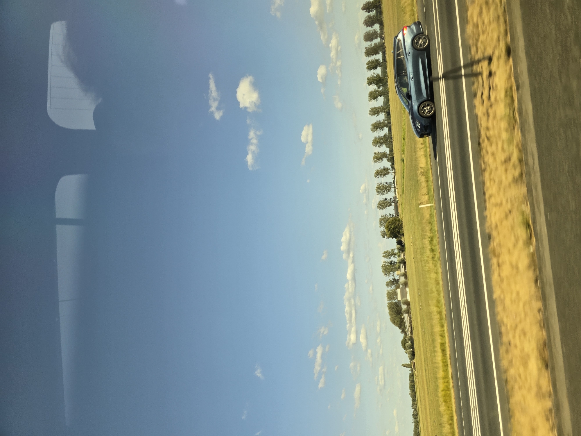 The image depicts a blue station wagon driving on a road with multiple lanes in Beuningen, Netherlands. The car is positioned in the right lane, moving from left to right. The road is bordered by a grassy median and a field with dry, yellow grass on the left. In the background, a row of trees lines the edge of a green field, and a building complex is visible further in the distance. The sky is a clear blue with scattered white clouds. The photograph appears to have been taken from a moving vehicle, as evidenced by the motion blur in the foreground and the reflections on the surface it was captured through.