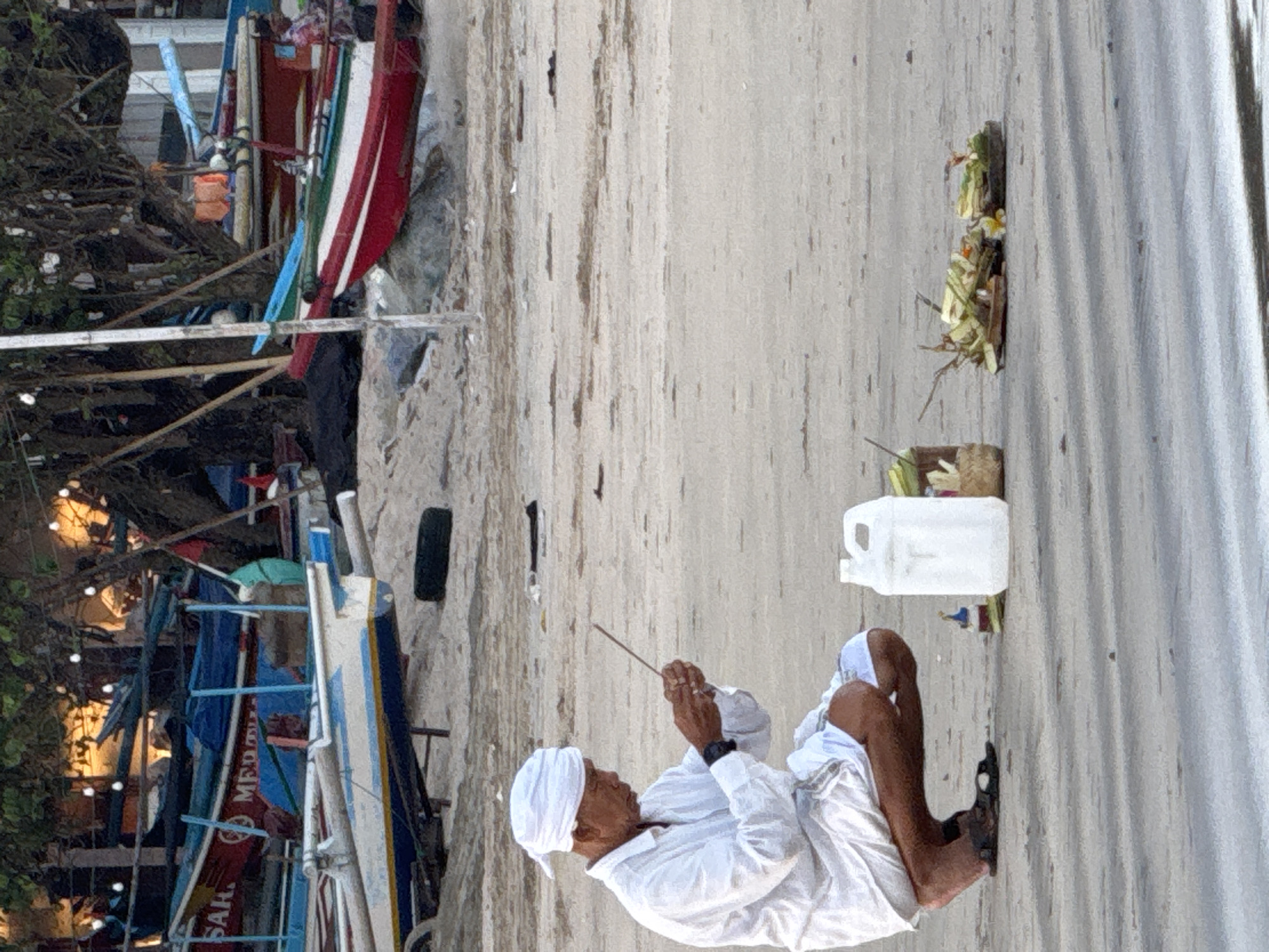 A man kneeling on a sandy beach in Jimbaran, Indonesia, is holding an incense stick. He is wearing a white shirt, pants, and a head covering. Next to him is a white plastic container and a small woven basket holding flowers and leaves. In the background are several boats. The beach surface shows a horizontal texture, possibly from being raked.