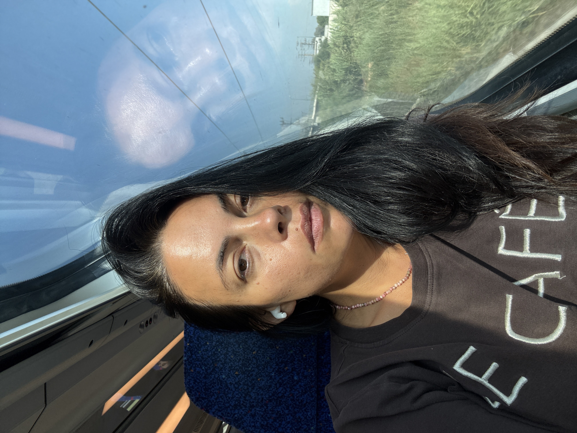 A dark-haired woman with fair skin is sitting on a train, looking forward and to her right. She is wearing a dark brown long-sleeved shirt with white lettering that reads "CAFE." She has a pink beaded necklace on and is wearing white wireless earbuds. Sunlight streams in from a large window to her right, reflecting on the window and showing a blurred image of the sky and some power lines. Outside the window, there is green vegetation and some structures that appear to be in an industrial or suburban area. The train's interior is visible behind her, with a dark blue patterned seat and overhead lighting. The image is a selfie, taken from the woman's perspective. The location is identified as the city of Petah Tikva, Israel.