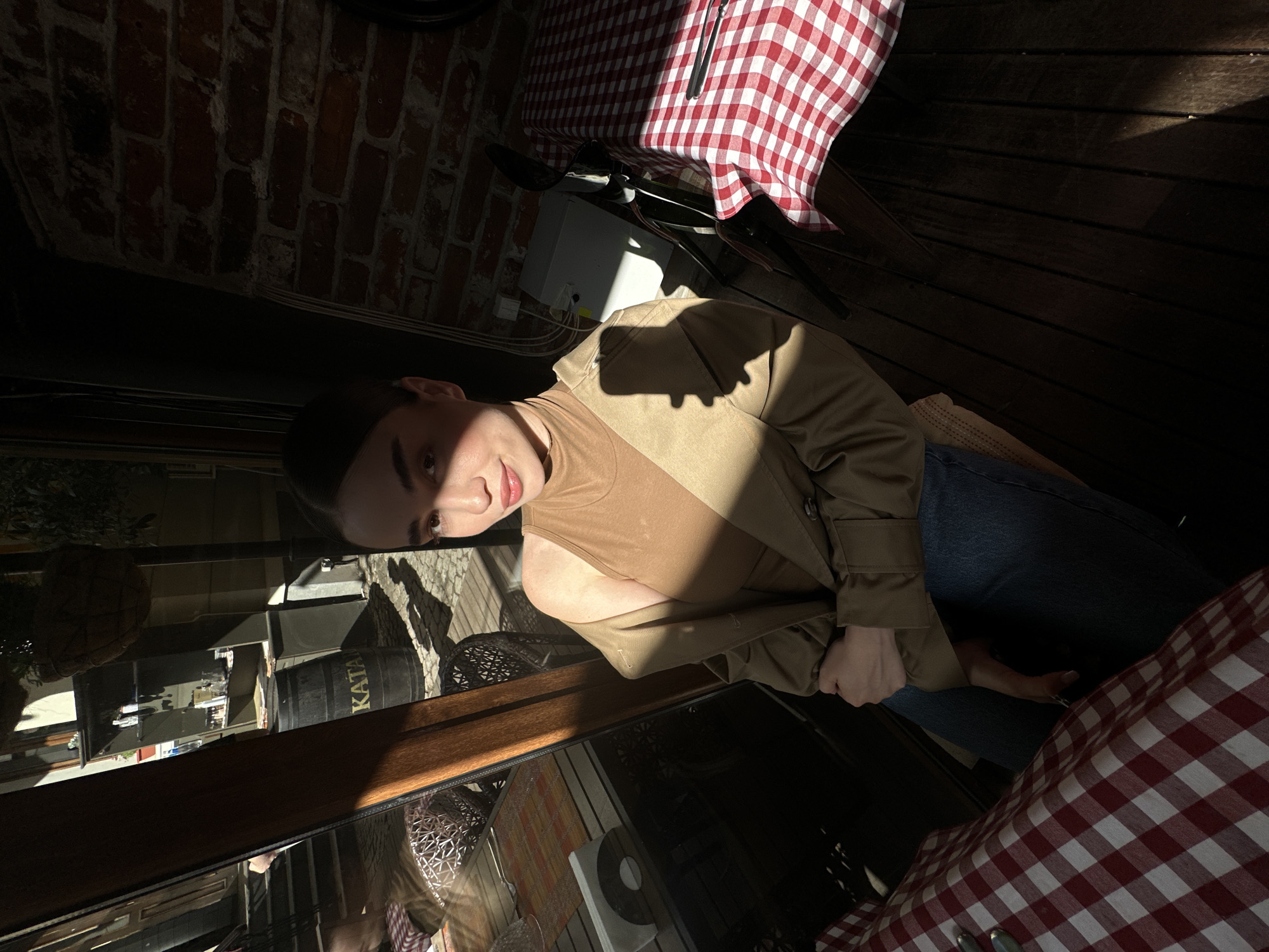 The image shows a woman seated at a table outdoors, presumably at a restaurant in Sofia, Bulgaria. She is wearing a light brown tank top and a khaki jacket, along with blue jeans. The table has a red and white checkered tablecloth. The environment suggests a semi-enclosed patio area with brick walls visible in the background. There is sunlight illuminating the scene, casting strong shadows. In the reflection on the window in front of the woman, there is a barrel marked "KATAN" and other elements of the city landscape. There is tableware on the table next to the tablecloth.