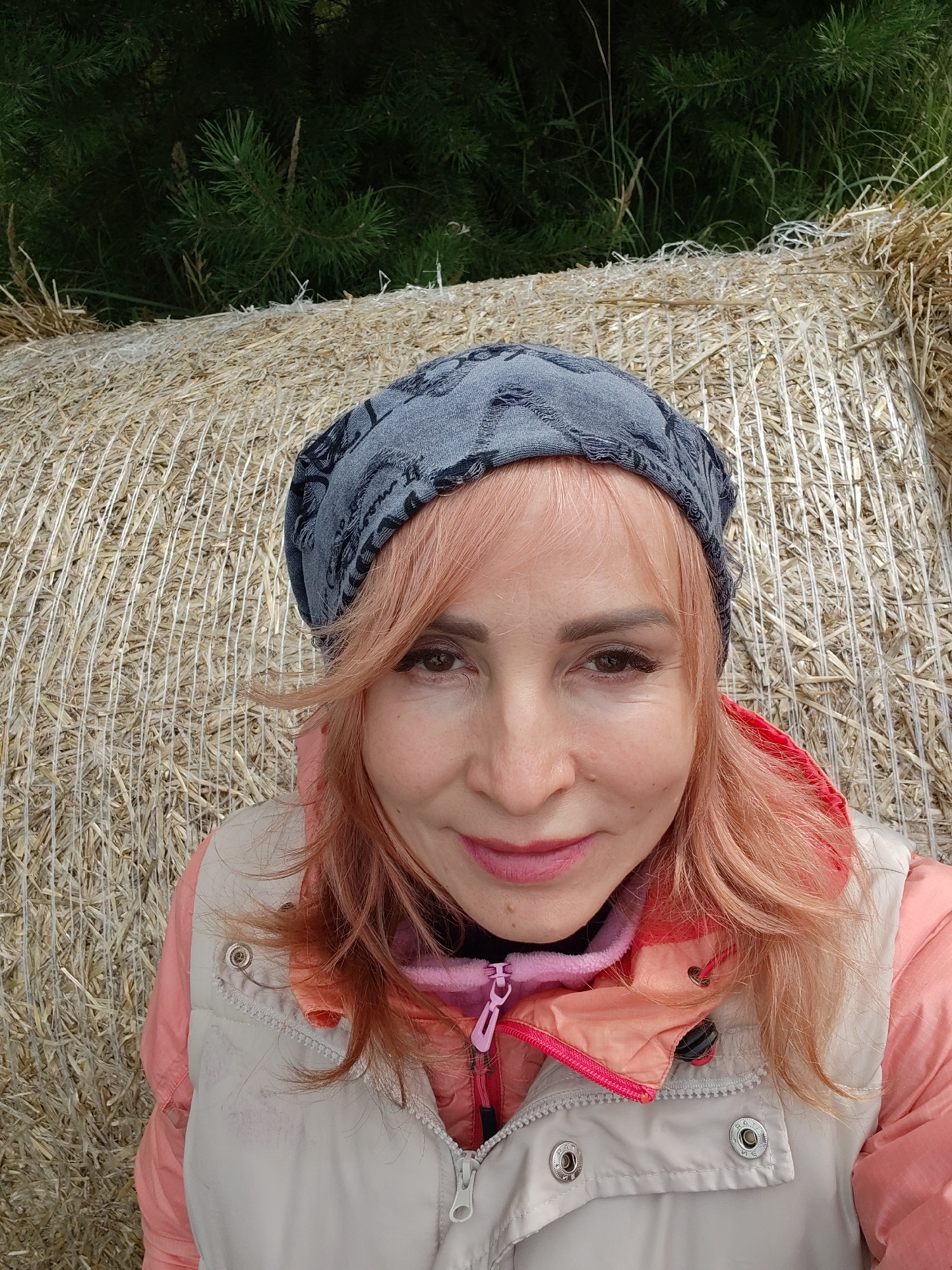 A medium shot photo shows a woman from the chest up. She is sitting in front of a large hay bale with green trees in the background. The woman has light skin, light brown eyes, and pinkish-blonde hair. She is wearing a grey patterned beanie, a light colored vest over a pink jacket, and a pink hood. The woman is looking directly at the camera and smiling slightly. The hay bale is tightly wrapped with white netting. The trees in the background are dark green and appear to be pine trees. The location is in Sidorovy Gory, Russia.