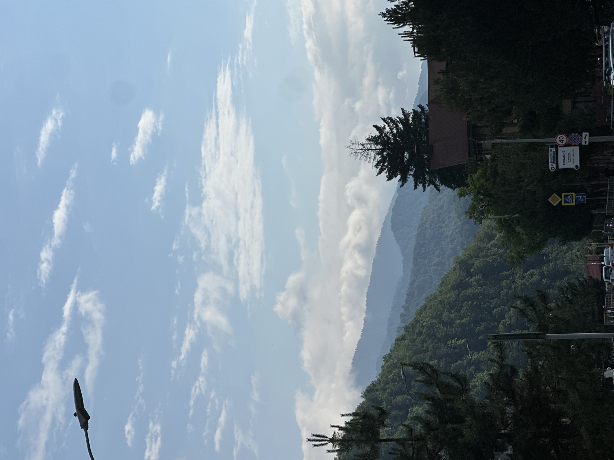 The image depicts a scenic view of rolling, verdant mountains under a bright, partly cloudy sky. The foreground is dominated by the silhouettes of trees and a streetlamp. In the mid-ground, a building with a brown roof is nestled among the trees, and a series of mountain ranges recede into the distance, their peaks softened by atmospheric haze. The sky is a pale blue, with scattered white clouds that create a sense of depth and movement.
A street sign is visible on the right side of the image. It has Cyrillic text and includes a circular sign with the number "40" within it, indicating a speed limit. Another sign below it displays "СВОБОДНО" (which translates to "Free" or "Available" in Russian) and a phone number.
The overall impression is one of a peaceful, natural setting, likely during the daytime. The weather appears to be fair, with sunlight filtering through the clouds. The presence of the streetlamp and signs suggests a road or inhabited area on the edge of the wilderness. The mountains, lush with trees, evoke a sense of a rural or semi-rural location. The orientation of the image suggests it might have been taken from a vehicle or a sloped surface.