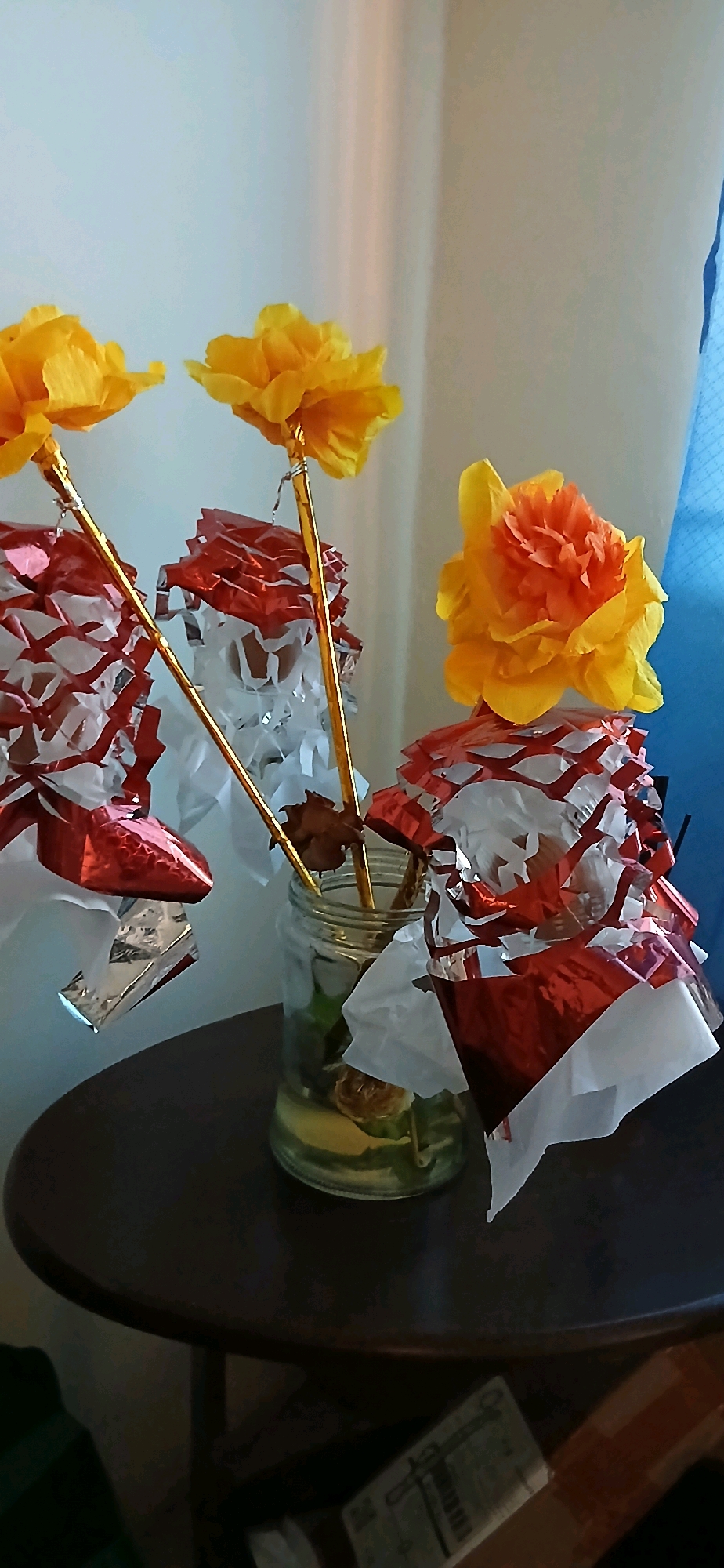 In Samarinda, Indonesia, a still life composition features three artificial flowers with yellow and orange petals on golden stems, placed in a glass jar containing what appears to be water and slices of fruit. The flowers are adorned with festive red and white crinkled paper decorations. The scene is set on a dark, round table against a light-colored wall, with a hint of blue fabric in the background. The overall impression is one of a handmade, celebratory display, possibly for a party or event.