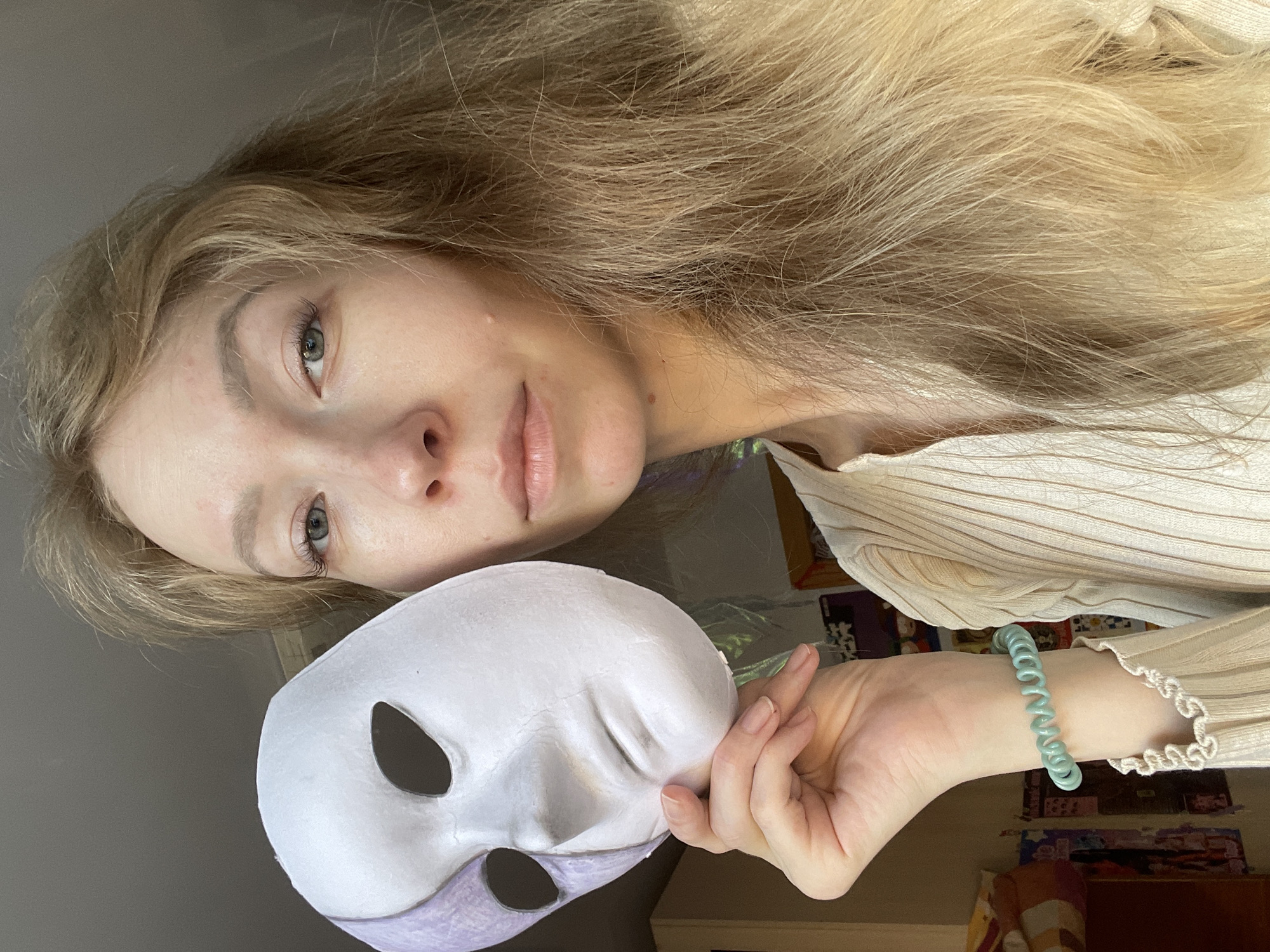 A young woman with long, blonde hair is holding a white mask in her right hand. She is looking directly at the camera. The mask has oval eye holes and a lavender section at the bottom. She is wearing a light-colored, ribbed shirt and a turquoise bracelet. The background includes a wall with posters and a wooden desk. The location is unknown.