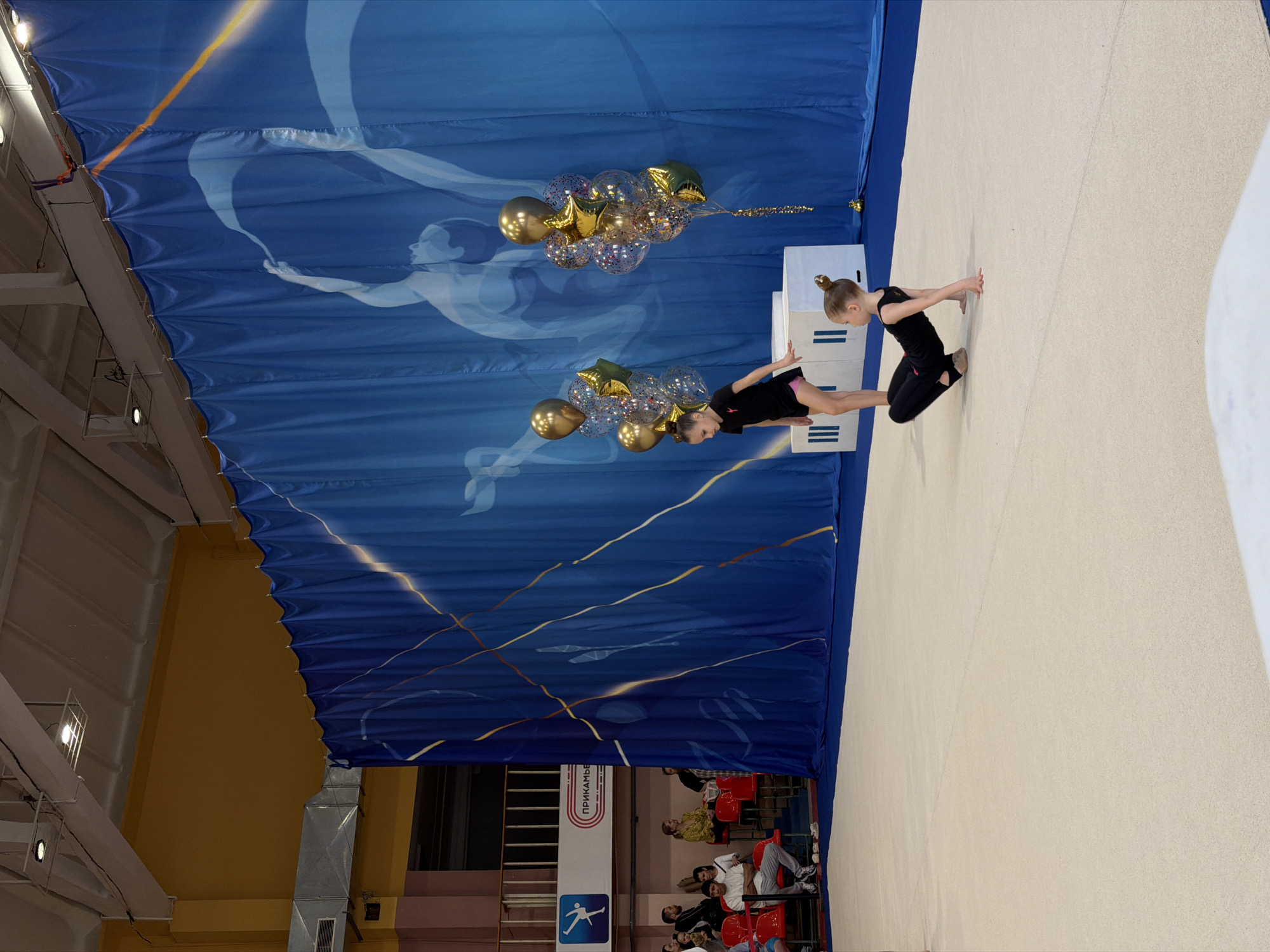 Two young gymnasts are performing on a light-colored mat in a gymnasium. The gymnast on the left is in a prone position on two stacked white boxes, while the gymnast on the right is kneeling on the mat, both with their hands touching the mat in front of them. Both are wearing black leotards and have their hair pulled back in buns.
A large blue banner hangs in the background, adorned with a silver image of a figure and abstract gold lines. Attached to the banner are two clusters of gold and clear balloons, with gold star-shaped balloons interspersed.
The gymnasium setting is evident from the metal framework, ventilation ducts, and a partial view of seating with spectators in the background. A sign with the inscription "ПРИКАМЬЕ" is partially visible. The gymnasium is likely located in Perm, Russia, based on the Cyrillic inscription.