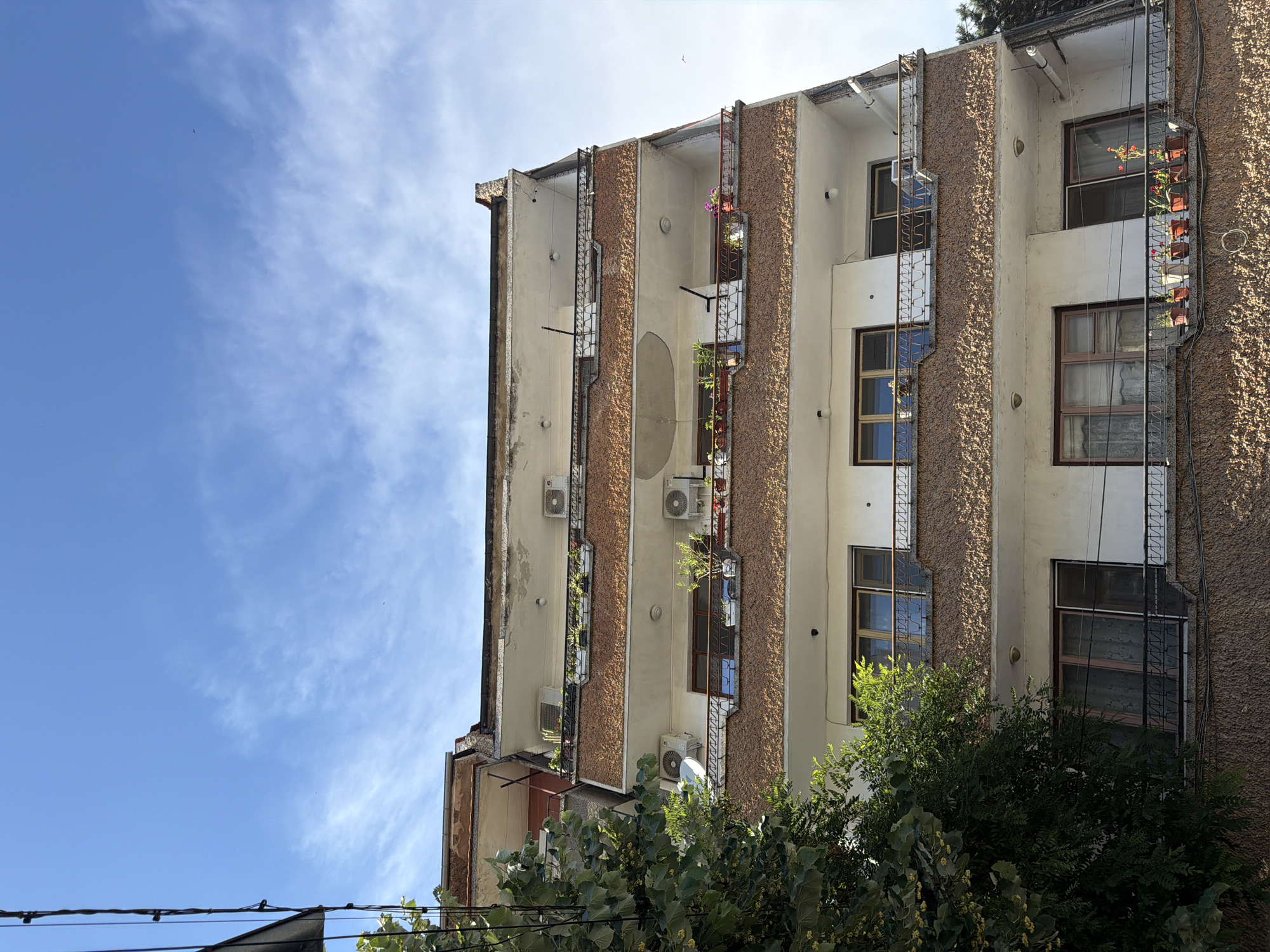 This is a ground-level, eye-level, medium shot of a building in Pazardzhik, Bulgaria. The building is a multi-story apartment complex with balconies on each floor. The facade of the building is white with brown textured panels on the balconies. There are several air conditioning units mounted on the exterior walls. Plants are visible on the balconies in planters. The sky is blue with some wispy clouds. A tree is visible on the left side of the image, with its branches extending into the frame. Electrical wires are also visible running across the top left of the image. The building appears to be in a residential area.