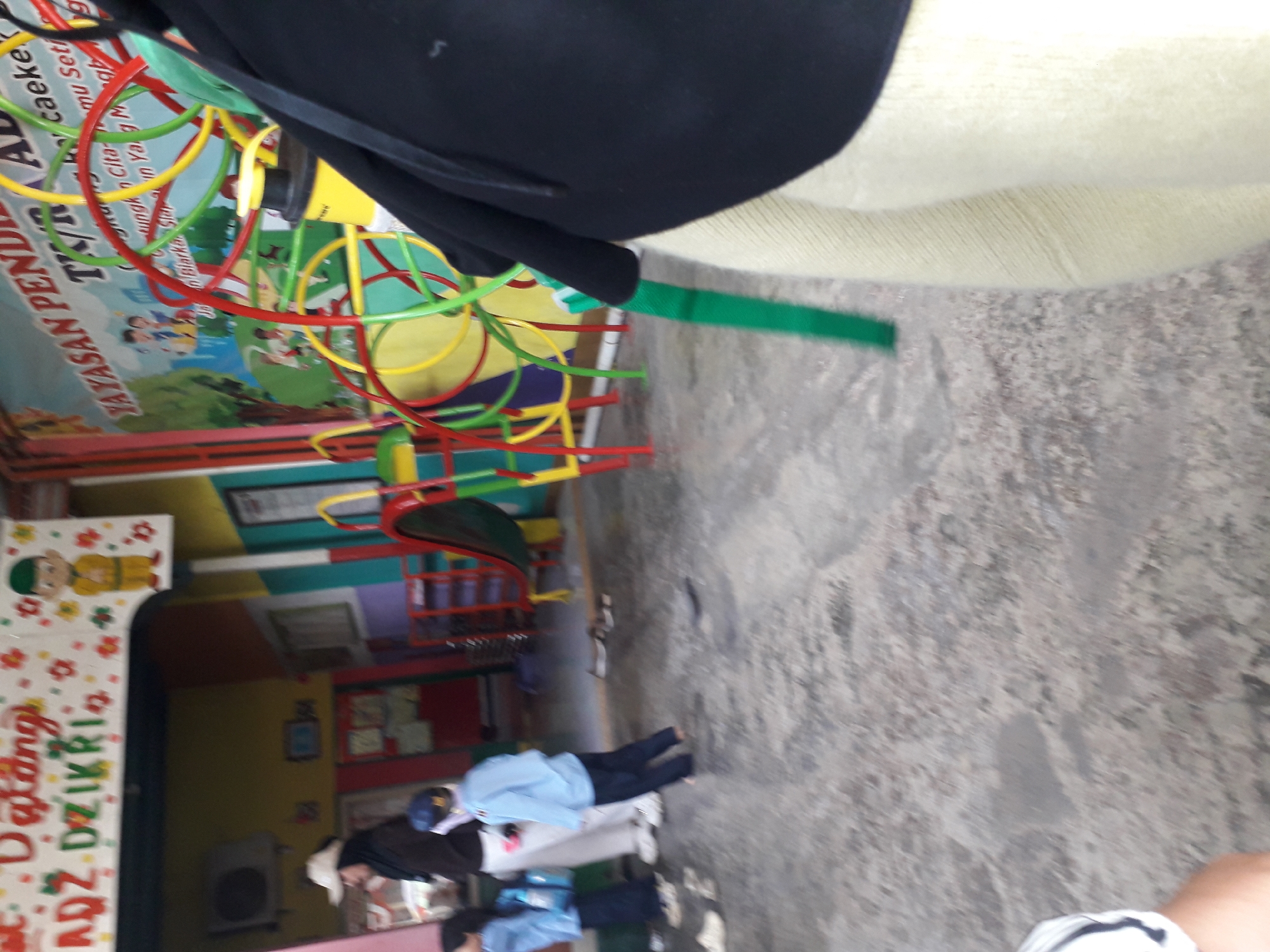 The image depicts an outdoor scene at what appears to be a playground or educational facility. In the background, a colorful mural is visible with text that includes "YAYASAN PEND" and potentially more Indonesian text. A spiral toy structure, comprised of red, yellow, and green loops, is also prominent. To the left, a woman wearing a white cap and a dark hijab stands near a wall adorned with a cartoon character and floral designs. Children in light blue uniforms are present, with one child walking away from the camera towards the right. The ground is a paved surface with visible wet patches. The overall setting suggests a public space, possibly a school or kindergarten, in Cankuang, Indonesia, as indicated by the text on the mural and the general atmosphere.
