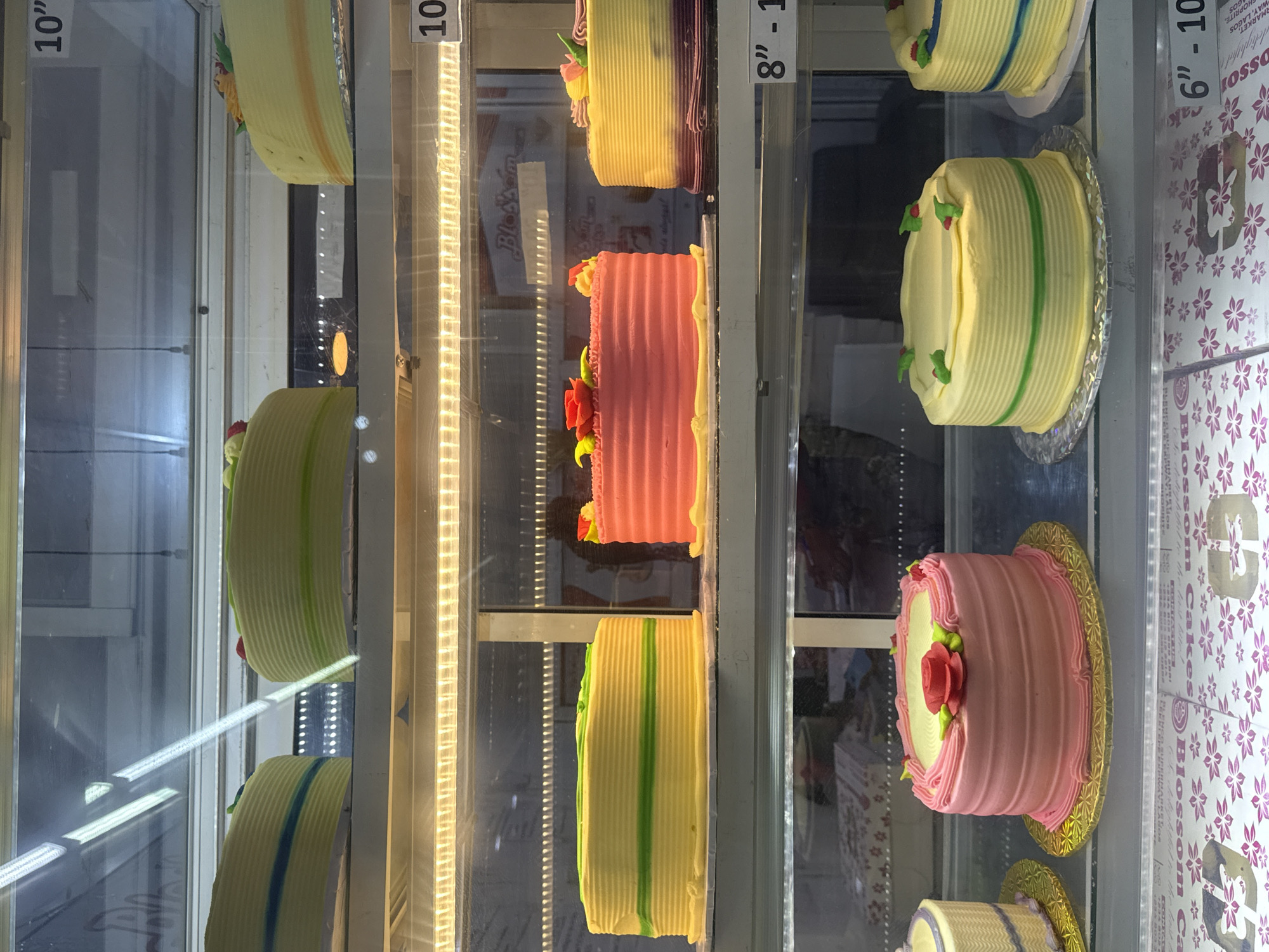 This is a close-up shot inside what appears to be a display case in a bakery. The case holds various cakes, primarily decorated with frosting in shades of yellow, green, pink, and purple. Many cakes have floral decorations. Signage indicates the sizes (6"-10", 8"-10", 10") and includes the name "Blossom Cakes", along with "MART, WAY, LAGOS", which, along with "NIGERIA, ADO", suggests a possible location. The scene is brightly lit.