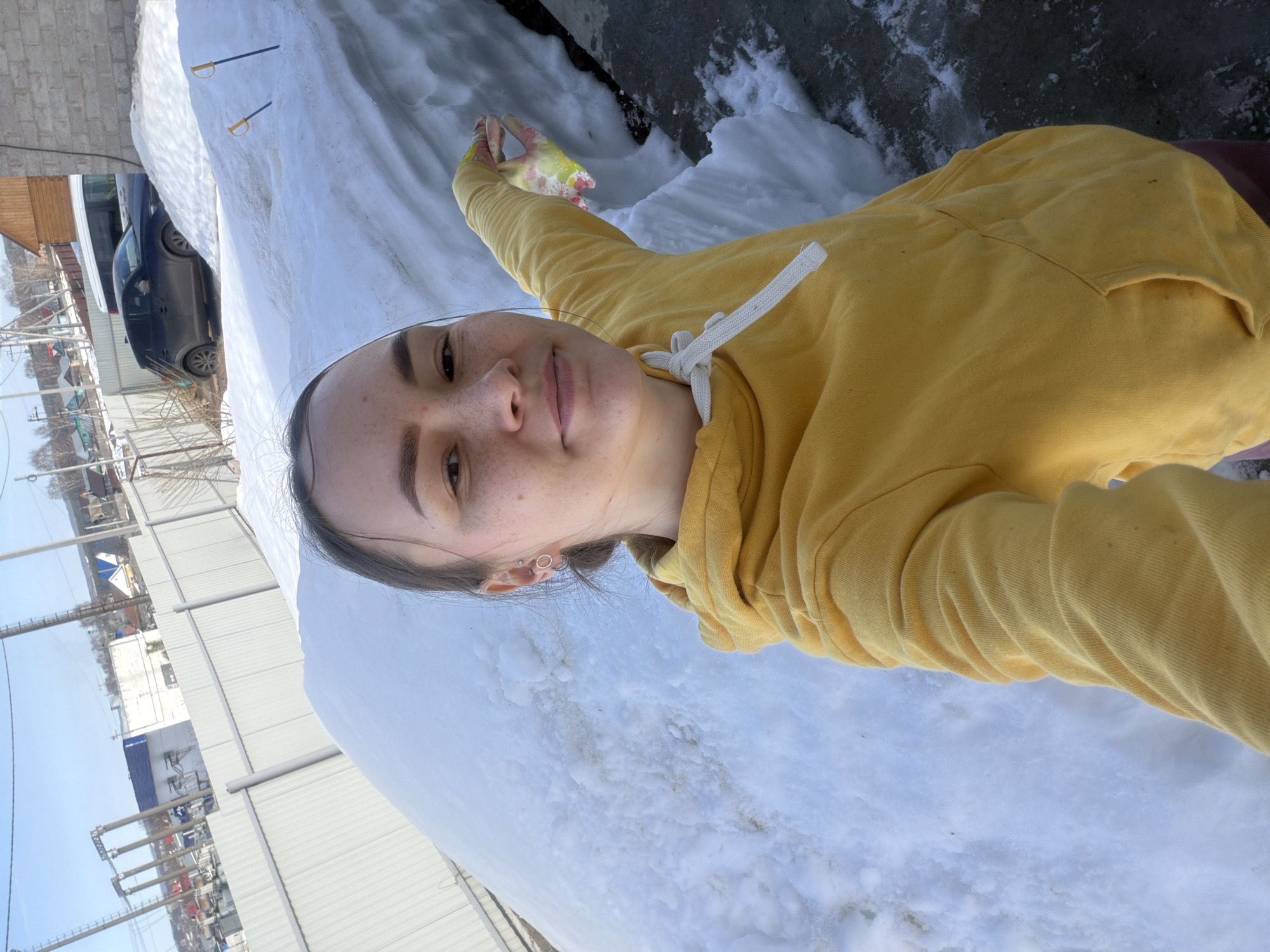 The image depicts a young adult female, characterized by a light complexion and visible facial freckles, with dark hair pulled back. She is wearing a yellow hooded sweatshirt with white drawstrings and displays multiple piercings in her left ear. Her right arm is extended, wearing a patterned glove with yellow, green, and pink floral designs, seemingly gesturing towards the environment while taking a selfie.
The immediate ground beneath her is dark concrete or asphalt, showing patches of melting snow and ice. To her left and behind her, a substantial, dirty snowdrift occupies a significant portion of the scene. Two yellow-handled ski poles are partially embedded in the snowdrift on the right side.
In the background, a light-colored corrugated metal fence spans horizontally. Beyond this fence, an urban or suburban environment in Chaikovsky, Russia, is visible under a clear blue sky. This includes various buildings, notably a white structure with a blue roof, alongside other residential buildings, utility poles with power lines, and scattered bare trees. A dark-colored SUV (appearing similar to a Mazda CX-5) and another lighter-colored vehicle are parked among the background structures, partially obscured by snow and buildings. The scene indicates a late winter or early spring period, with significant snow present but clear skies and evidence of melting.