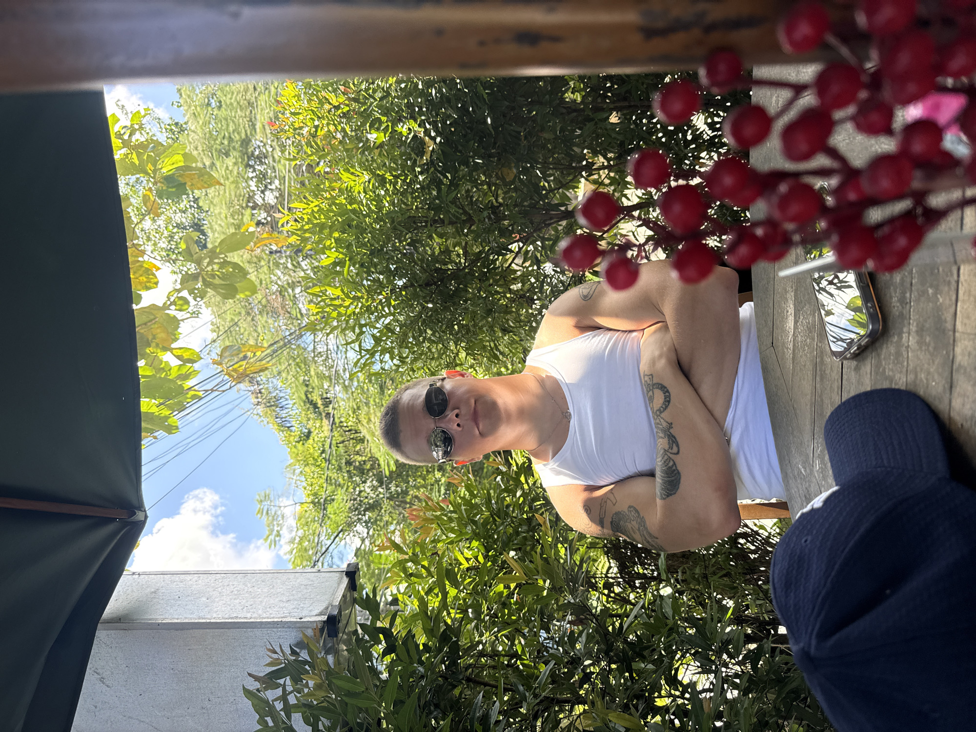 A photograph, taken in Kuta Selatan, Indonesia, shows a man with short hair wearing sunglasses and a white tank top, sitting at a wooden table. His arms are crossed. Tattoos are visible on his arms. A mobile phone rests on the table near a cluster of red berries. A dark blue baseball cap sits near the edge of the table. Green foliage surrounds the table, with glimpses of a building and blue sky visible through the leaves. An umbrella is visible in the foreground.
