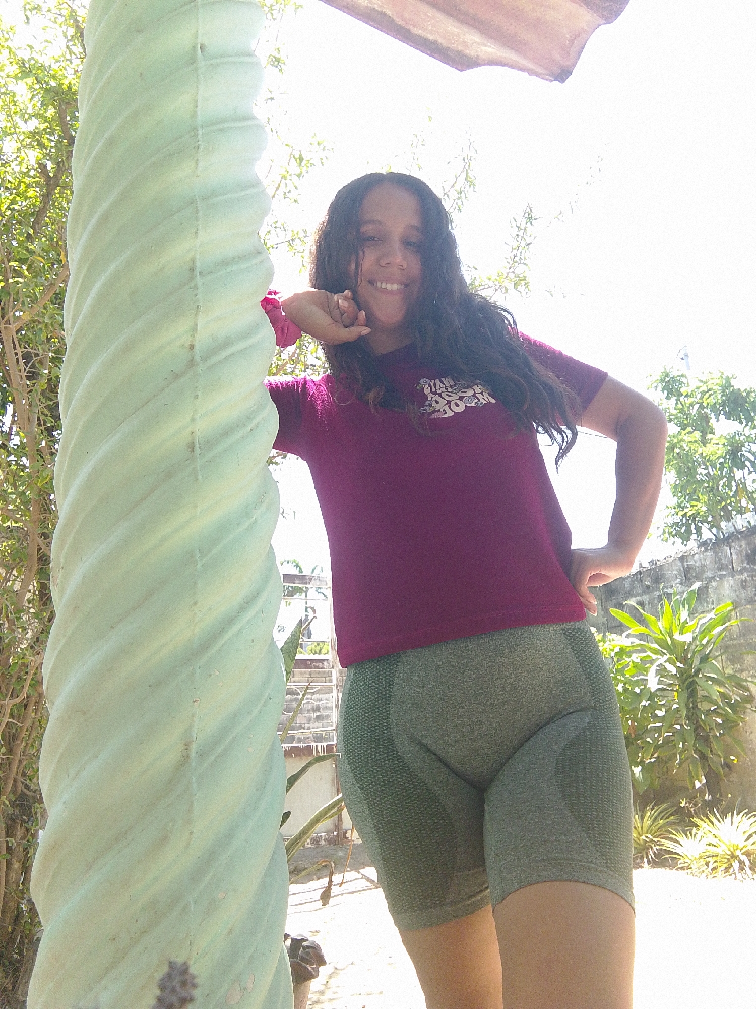 A woman with long, dark, wavy hair is posing outdoors in Juan Griego, Venezuela. She is wearing a maroon t-shirt and olive green athletic shorts. She is leaning against a tall, light green, fluted column-like structure with her left hand near her face and her right hand on her hip. The background includes trees, a wall, and other plants, bathed in bright sunlight.