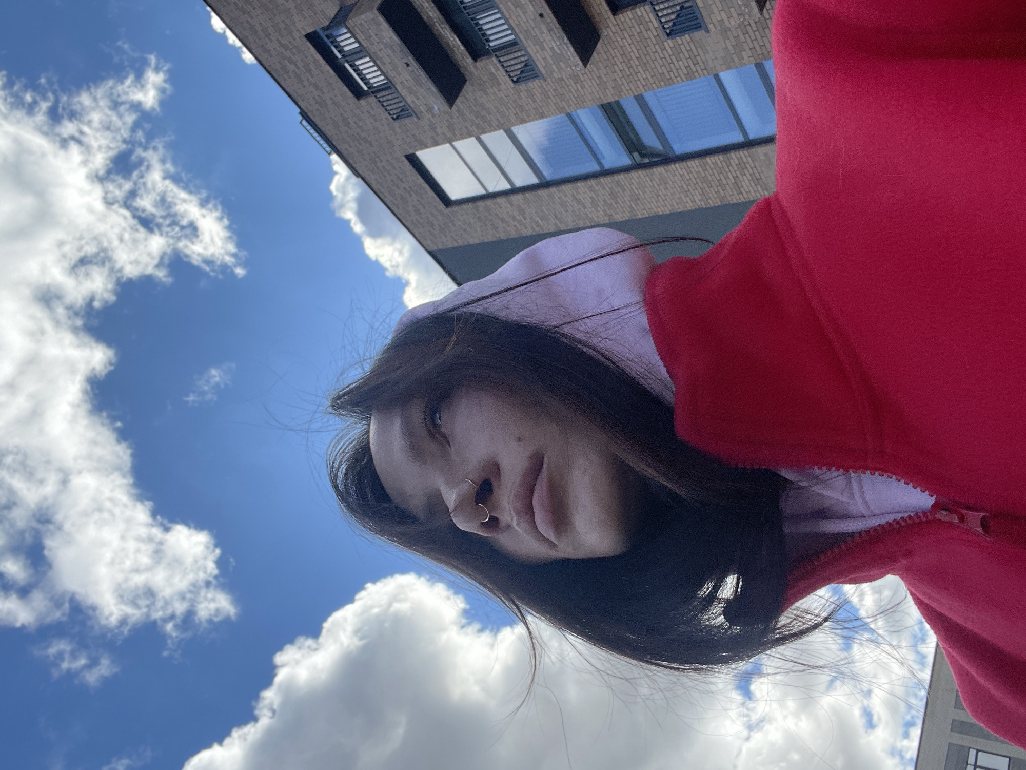 The image features a young woman seen from a low angle, looking downwards towards the camera. She has long dark hair and is wearing a red zippered hoodie over a lighter-colored hooded garment. She has a gold-colored hoop nose ring on her right nostril and a smaller stud on her left nostril. Her expression appears calm or neutral.
The background is dominated by a bright blue sky with scattered, fluffy white cumulus clouds, indicating good weather and daytime. Partially visible in the upper right quadrant of the frame is a modern building constructed of light brown or tan bricks, featuring large rectangular windows that reflect the sky. The perspective suggests the photograph was taken from below, looking up at the woman and the building. There is no visible text in the image. The scene is consistent with an outdoor setting, potentially in an urban or suburban area such as Семёново, Russia.