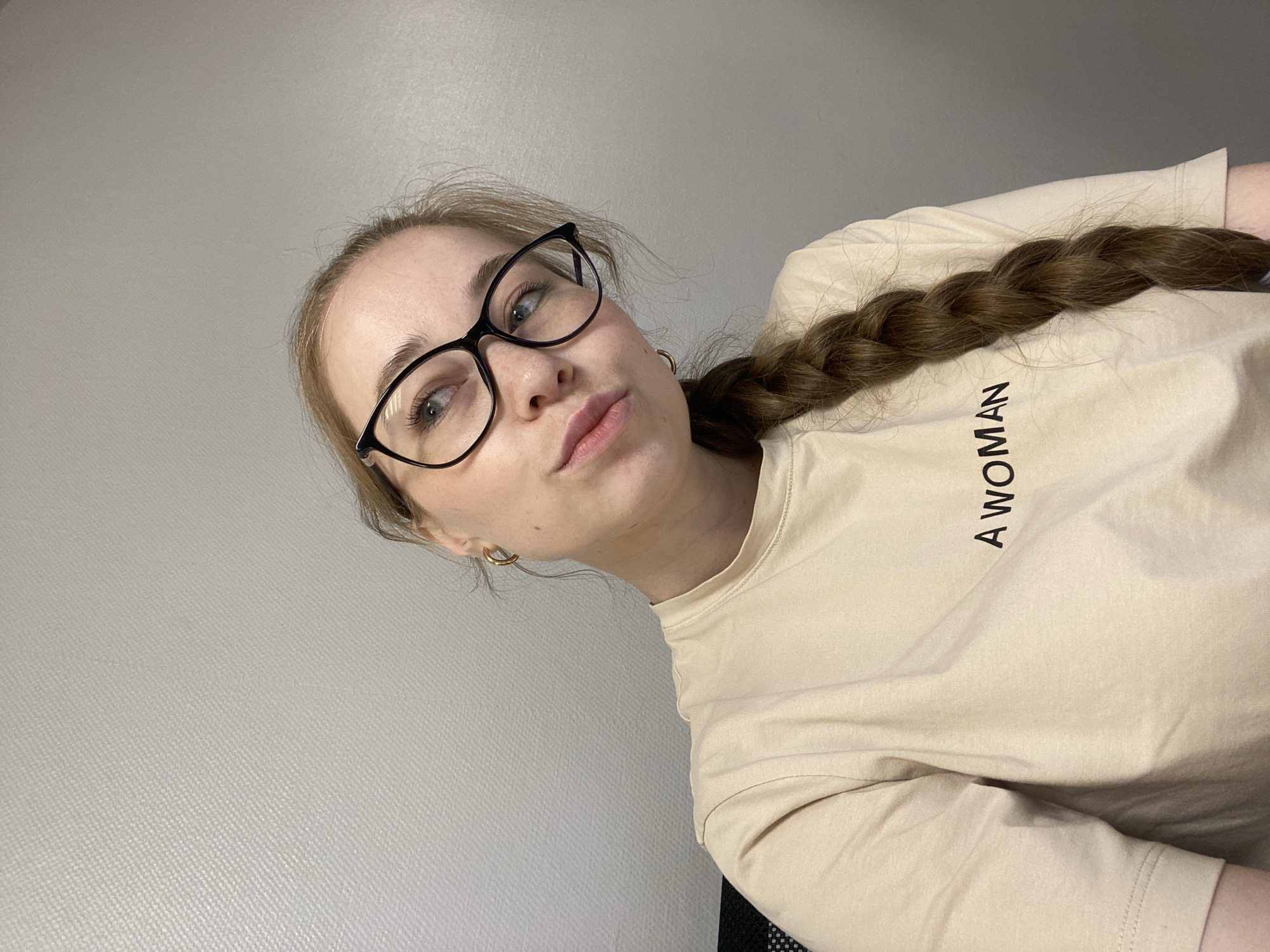 The image depicts a young woman indoors, captured from an angled perspective, likely a selfie. She has light brown hair styled in a thick braid resting over her shoulder. She wears black-framed eyeglasses, a light beige or cream-colored long-sleeved t-shirt with the text "A WOMAN" printed vertically in black letters on the front, and a small gold hoop earring in her visible ear. Her expression is subtle, with a slight pout on her lips, and she looks directly towards the camera. The background is a plain light grey wall, suggesting an indoor setting with artificial lighting. The context provided indicates the photo was taken in Saint Petersburg, Russia.